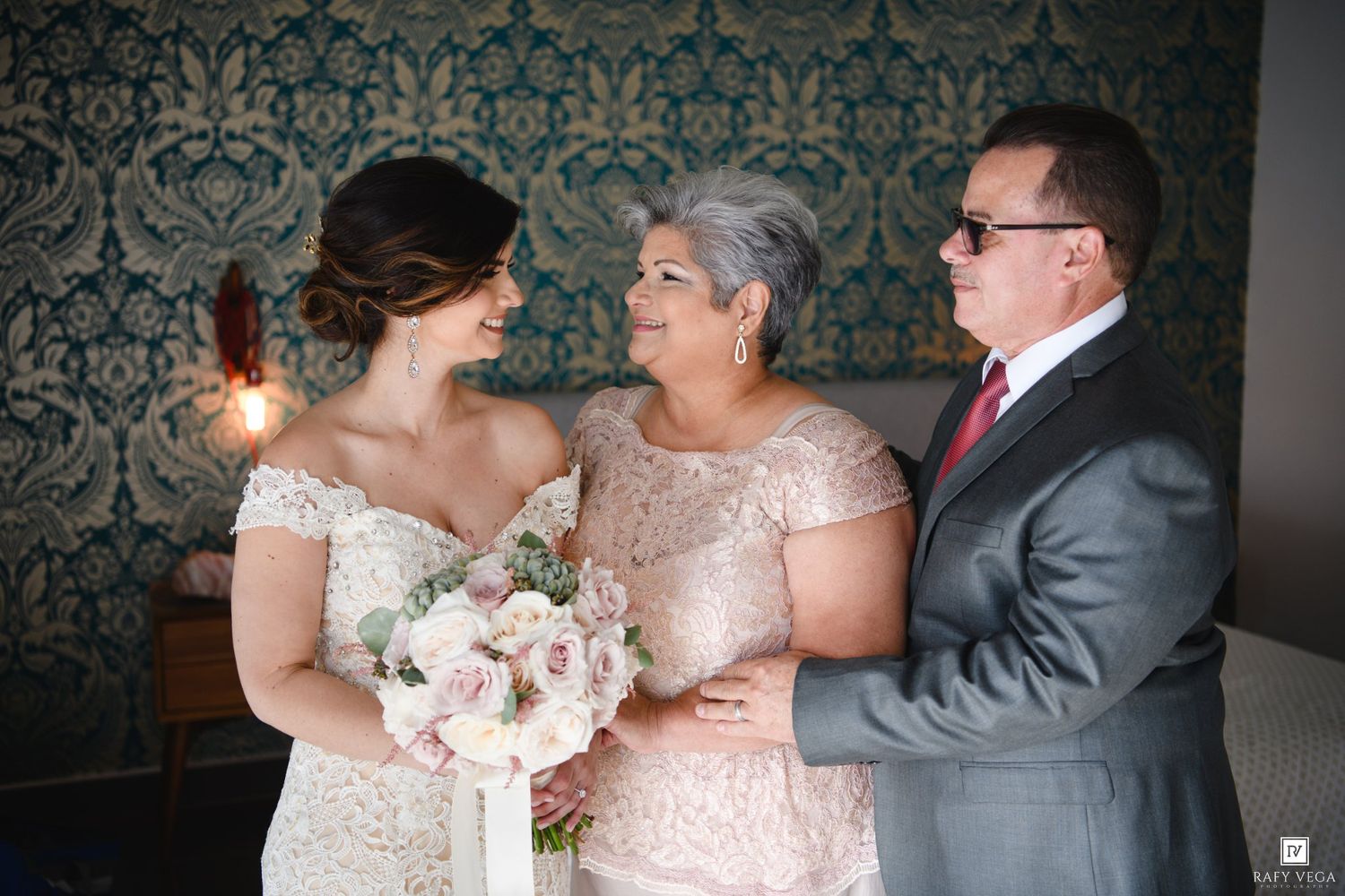 Bride in lace gown holds bouquet with mother in pink dress and father in gray suit at wedding.