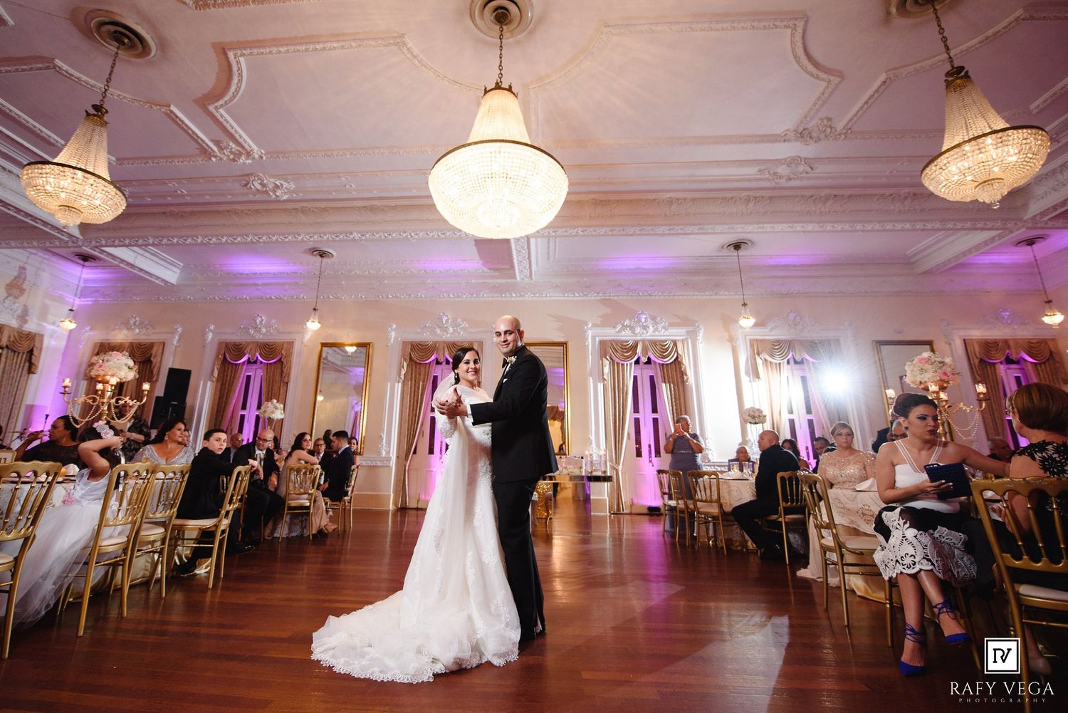 Bride and groom share first dance in elegant ballroom with gold chairs, chandeliers, and purple uplighting at wedding reception.