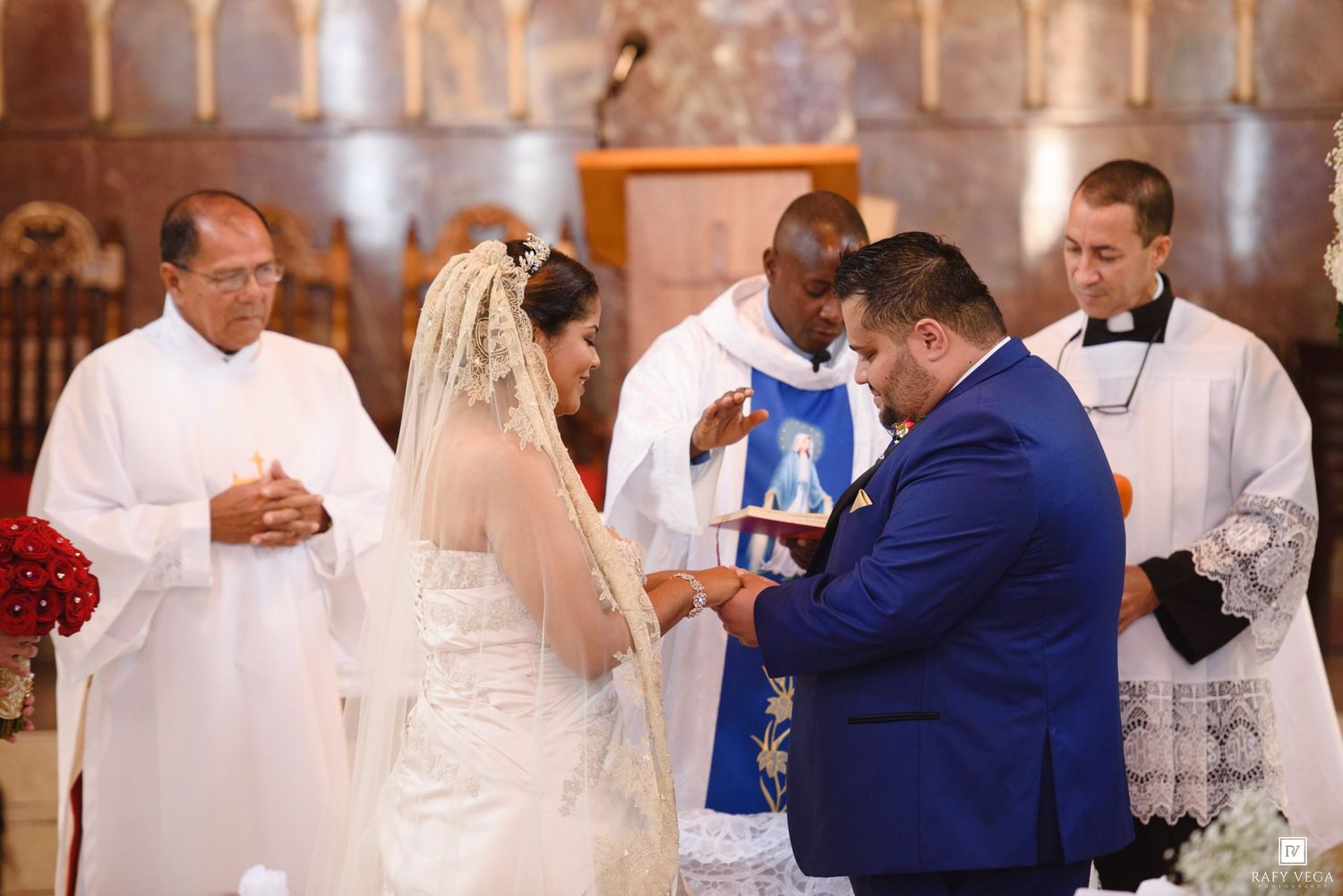 Bride and groom exchange rings during a Catholic church wedding ceremony with officiating priests in white robes.