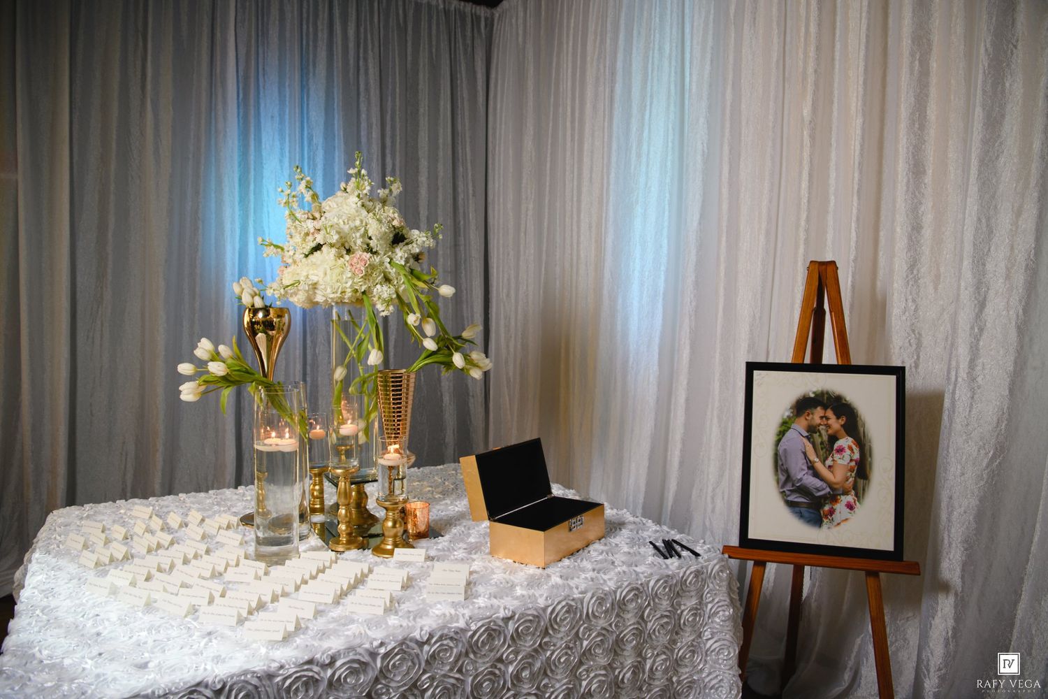 Elegant wedding display table with white floral arrangements, candles, and a framed couple portrait on a wooden easel.