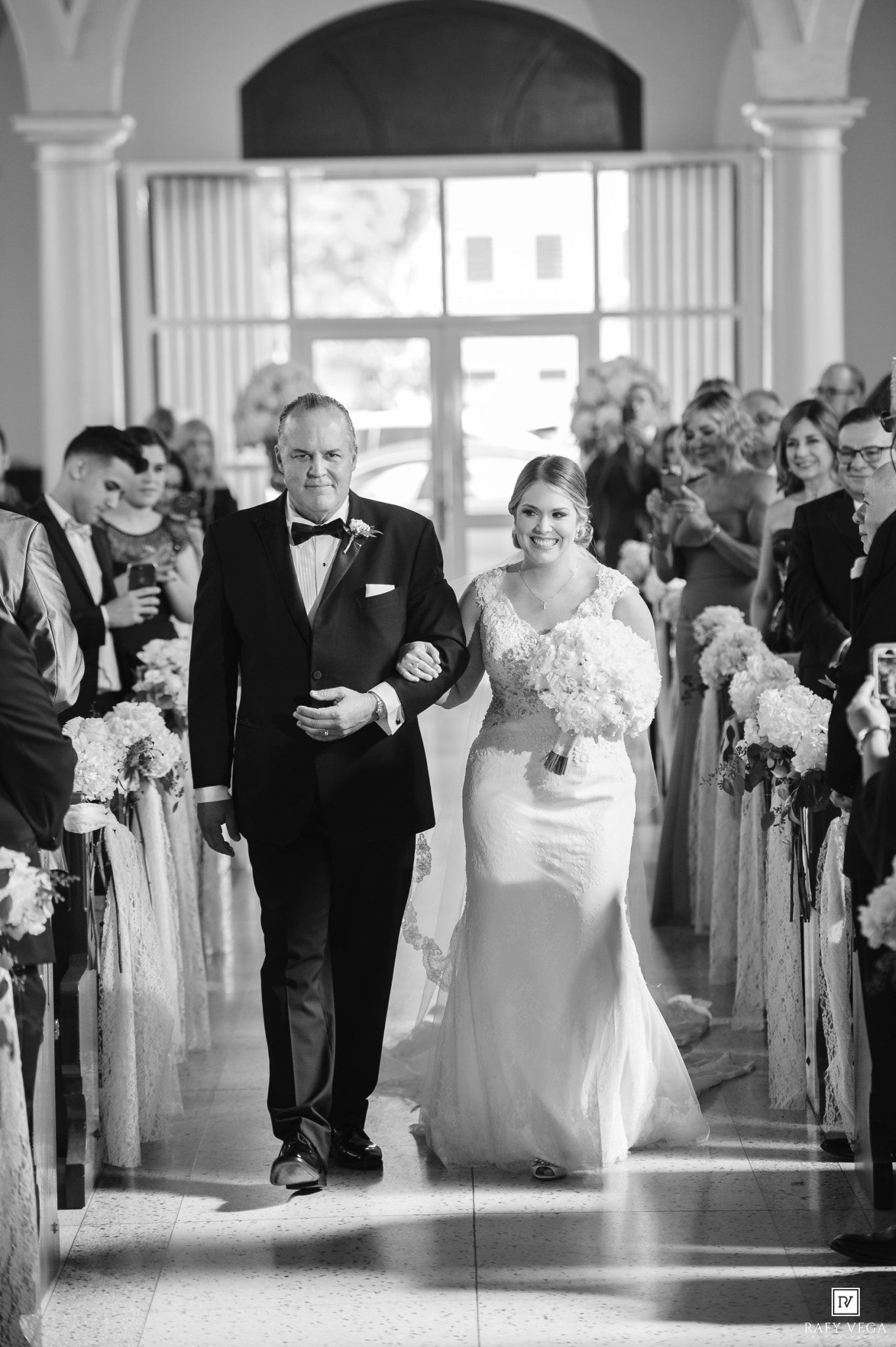 Bride walks down the aisle with her father in a classic black and white wedding ceremony photo.