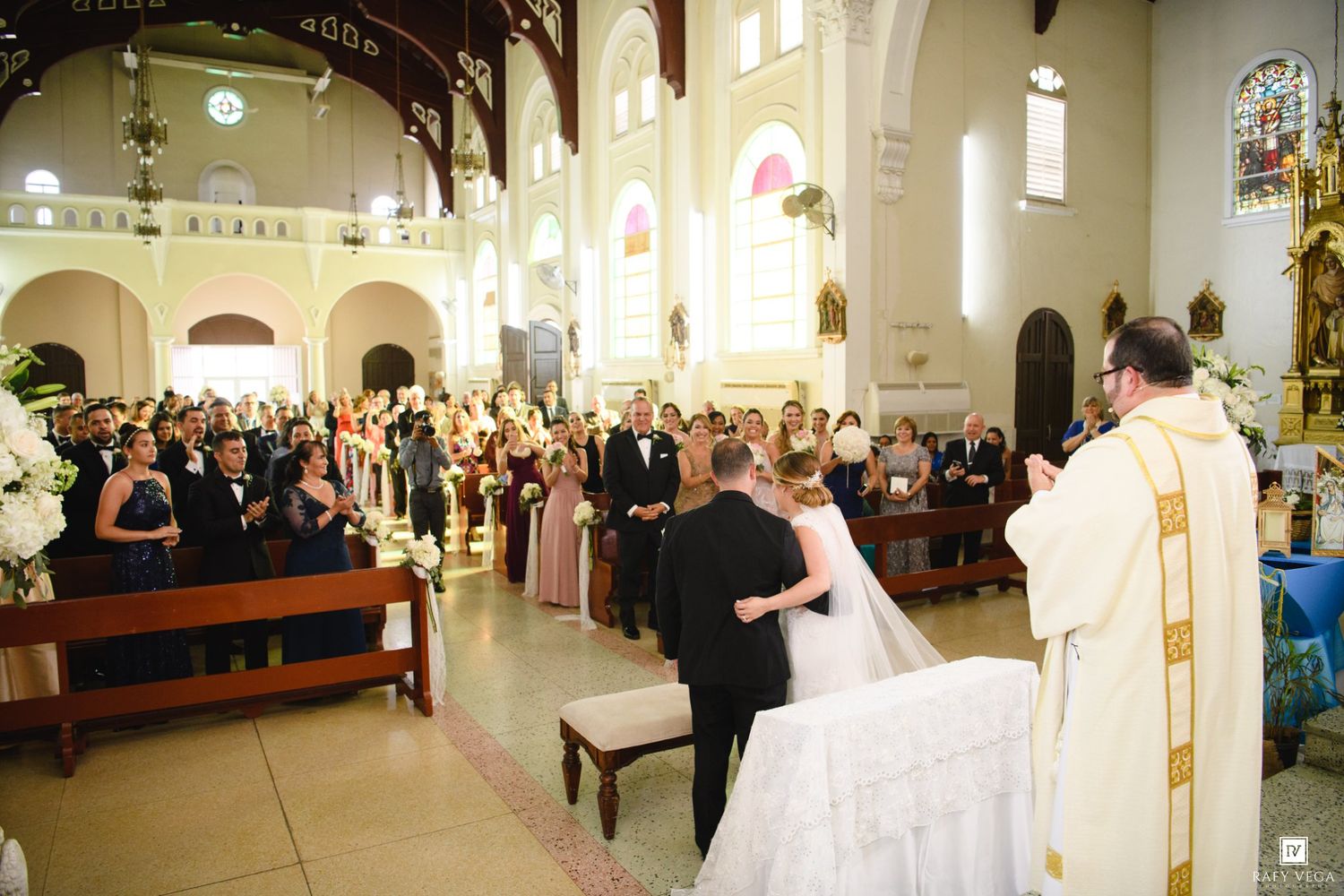 A bride and groom stand at the altar during a Catholic church wedding ceremony with guests seated in pews.