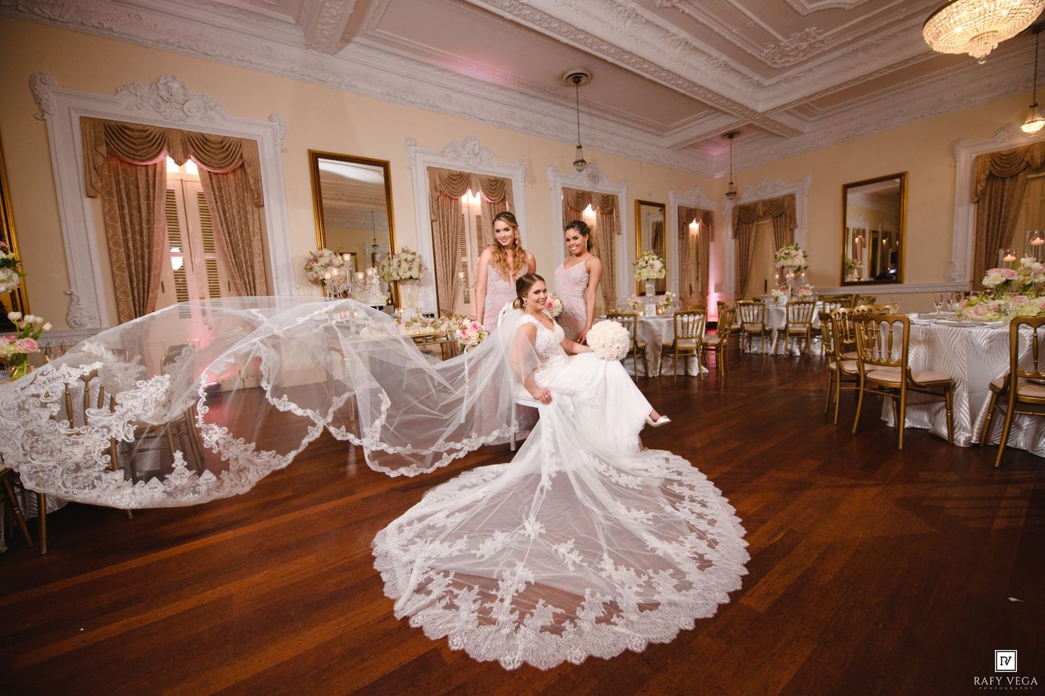 Bride in elegant lace wedding gown with flowing veil in ornate ballroom, bridesmaids in pink dresses nearby.