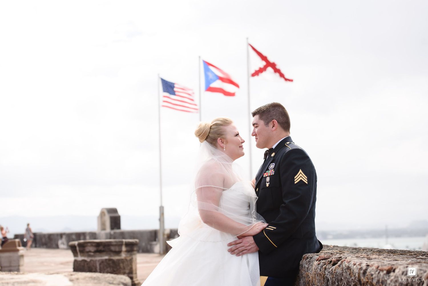 Military groom in dress uniform with bride in white wedding gown posing before American and Puerto Rico flags.