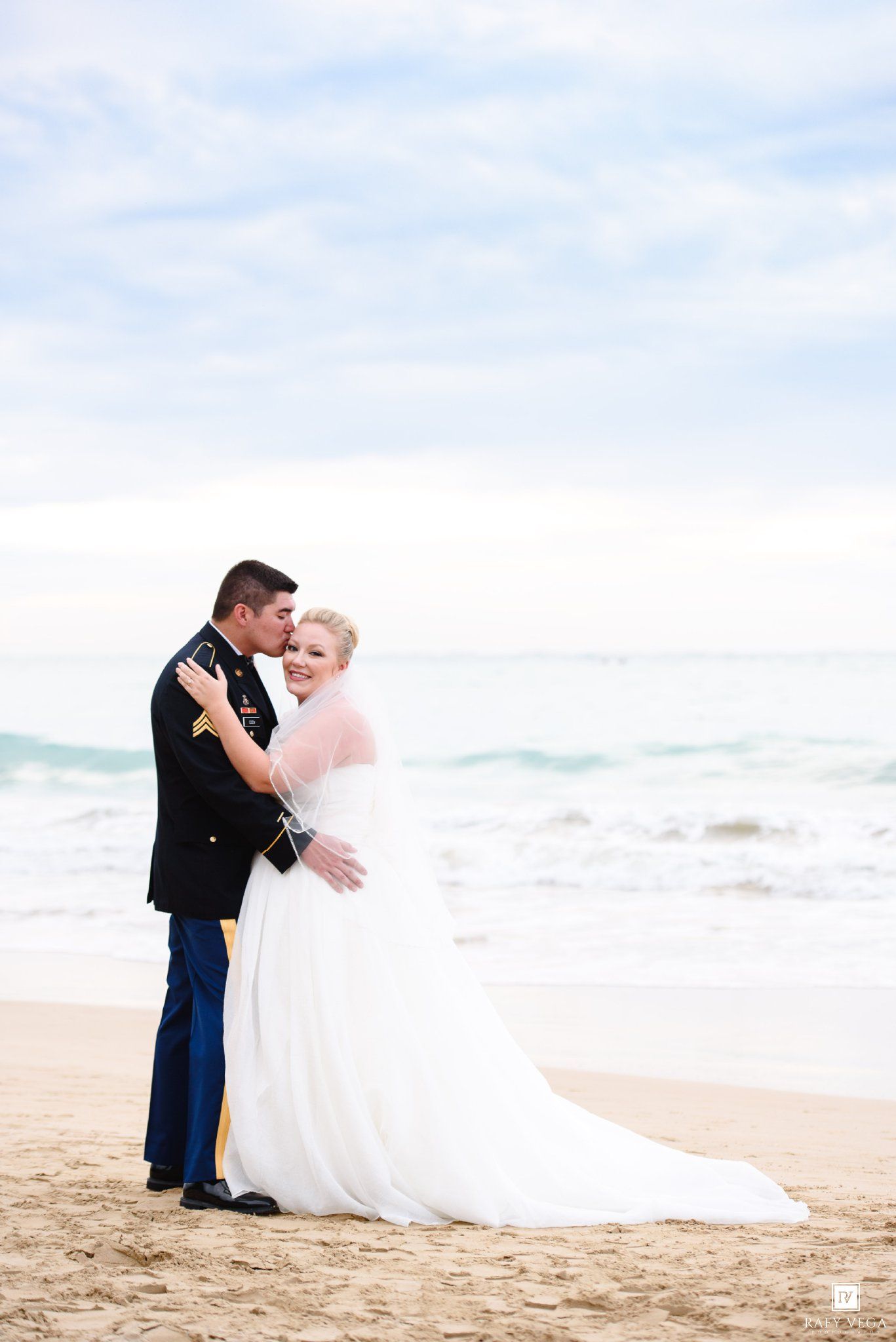 Military groom in dress uniform kisses bride in white gown and veil on sandy beach with ocean waves behind them.