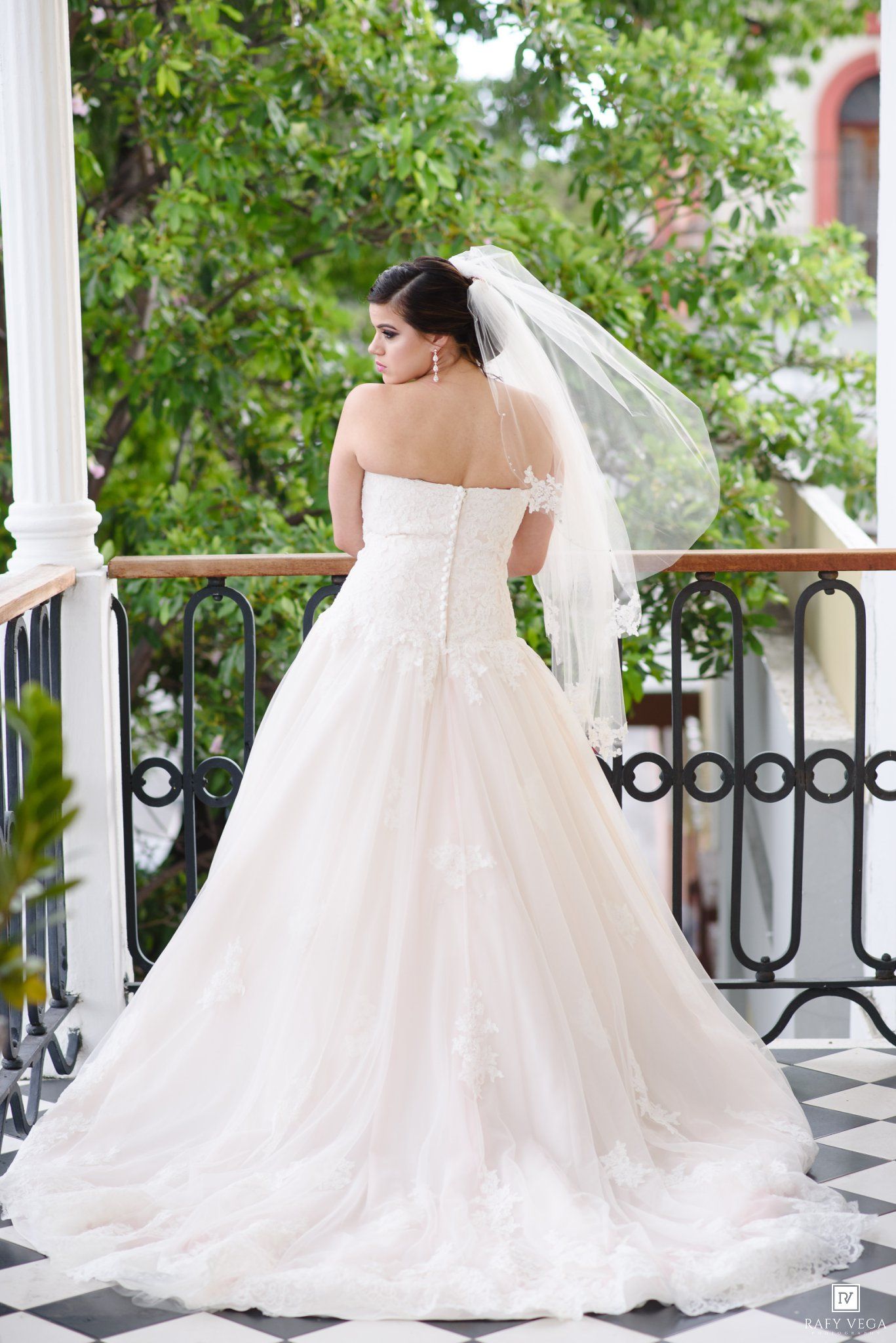 Bride in strapless lace ball gown with veil on balcony surrounded by lush greenery.