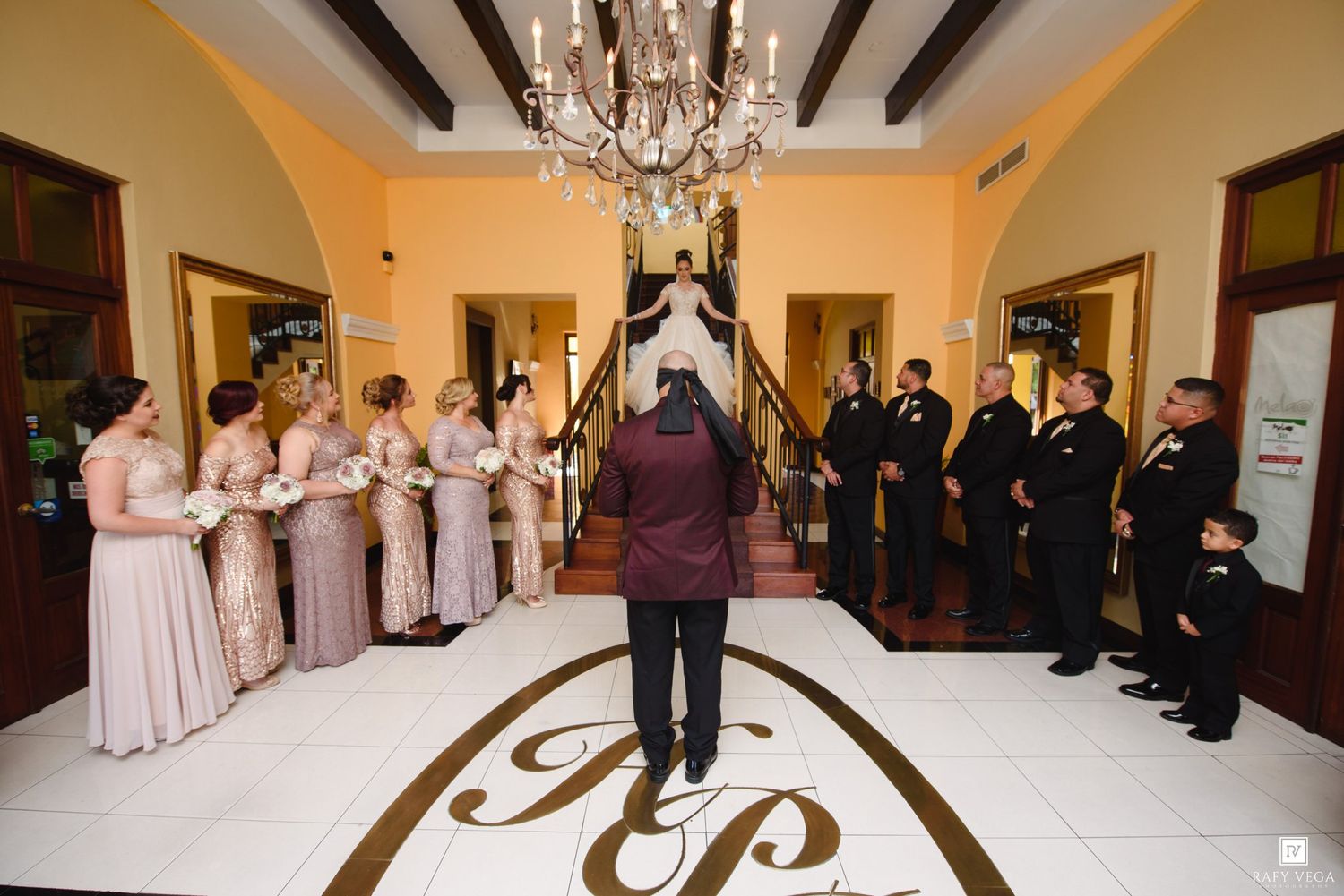 Bride descends staircase toward groom, flanked by bridesmaids in blush gowns and groomsmen in black suits, in elegant venue.