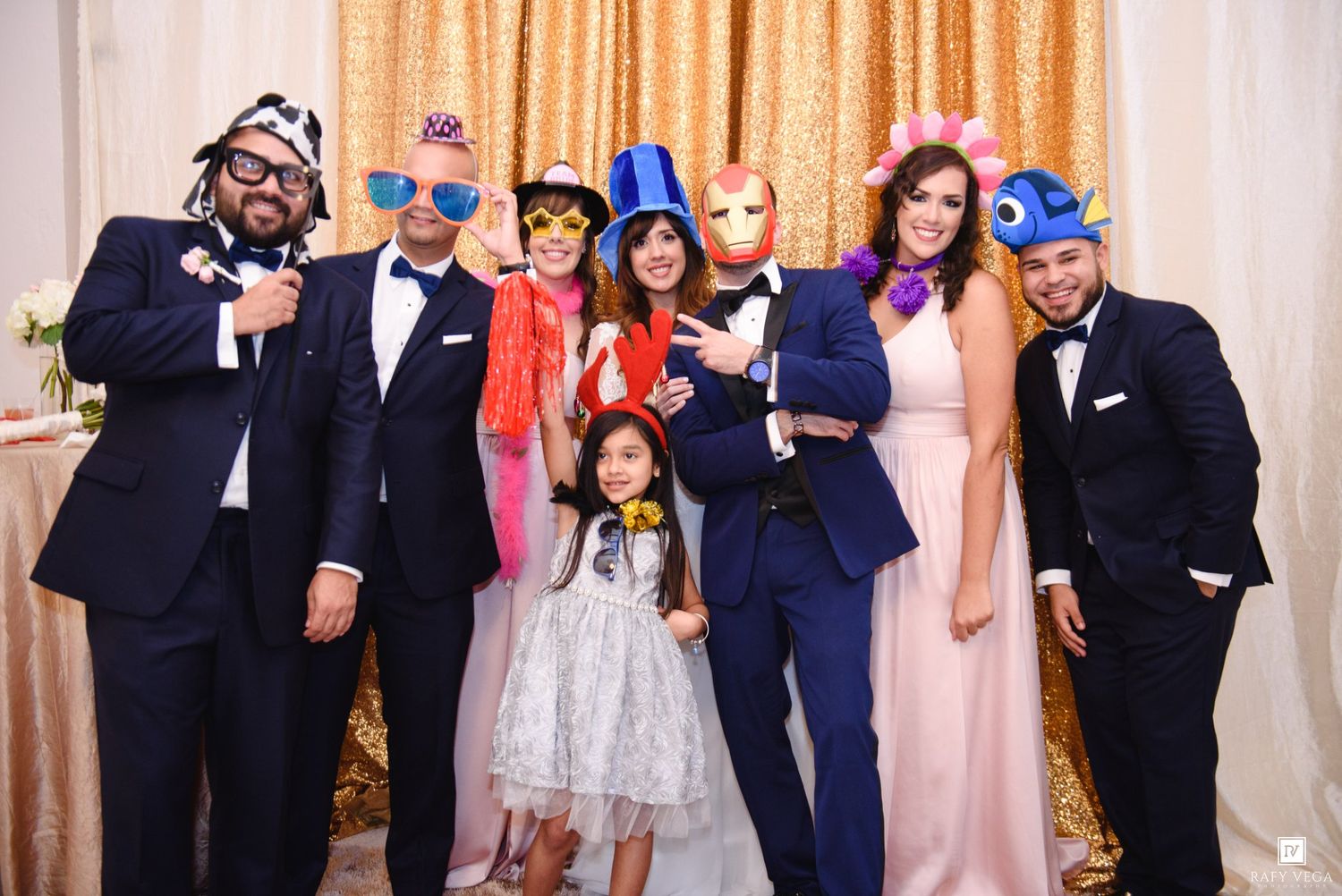 Group of seven adults and one child posing with colorful superhero masks at a formal event with gold backdrop.