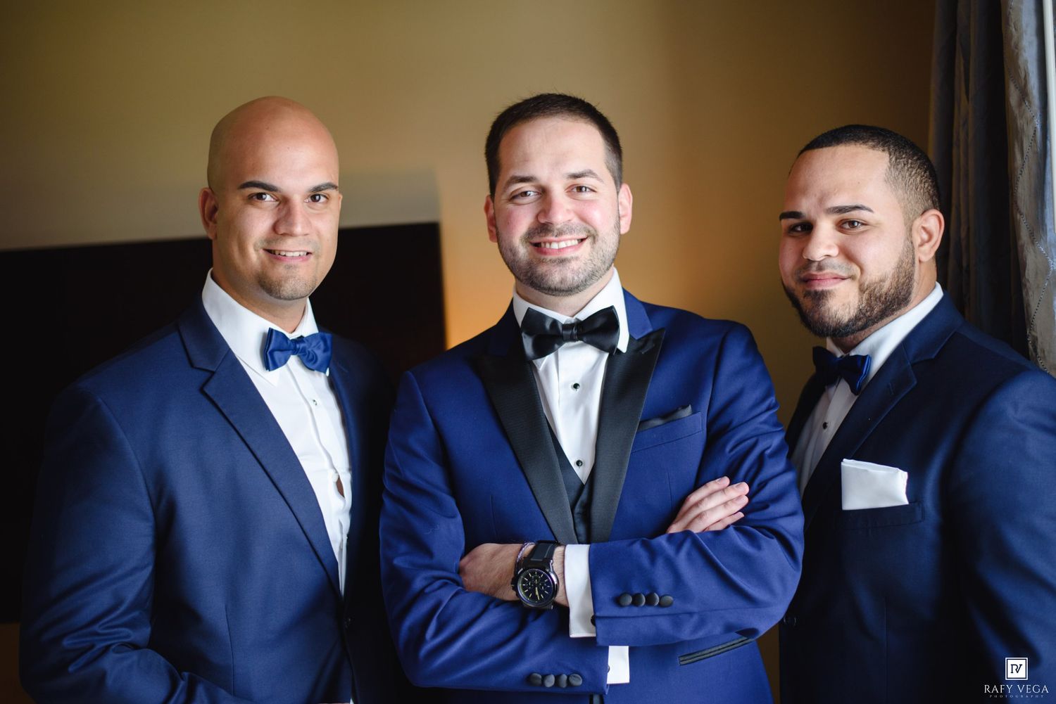 Three groomsmen in royal blue tuxedos with bow ties pose together smiling at a wedding celebration.