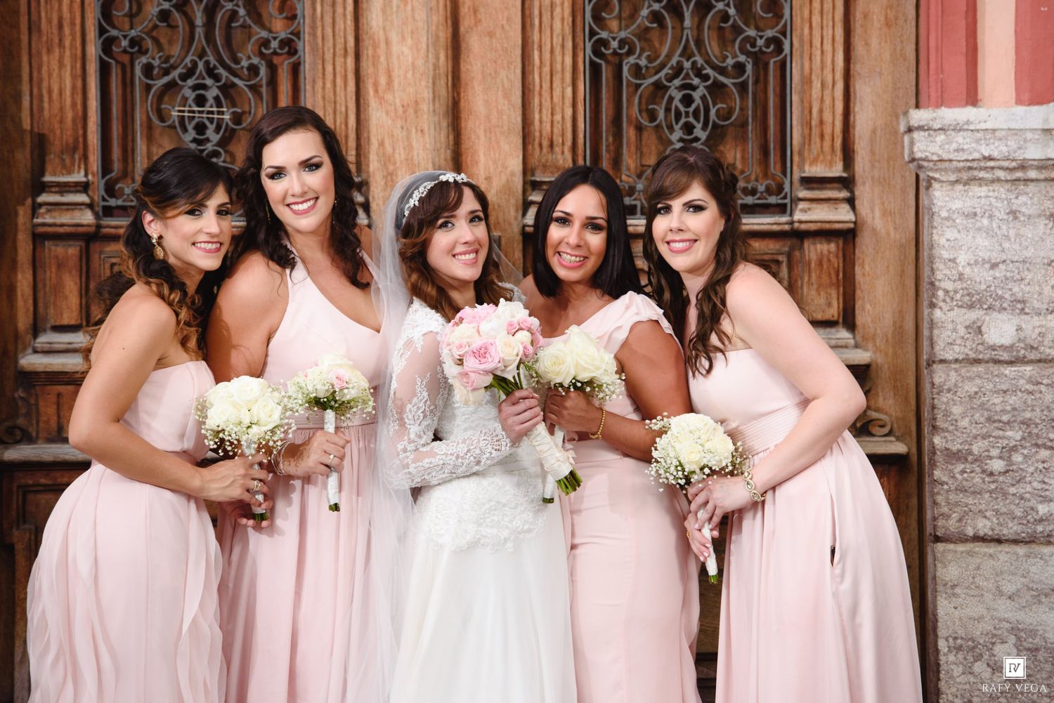 Bride in white lace gown with four bridesmaids in blush pink dresses holding bouquets outside ornate wooden doors.