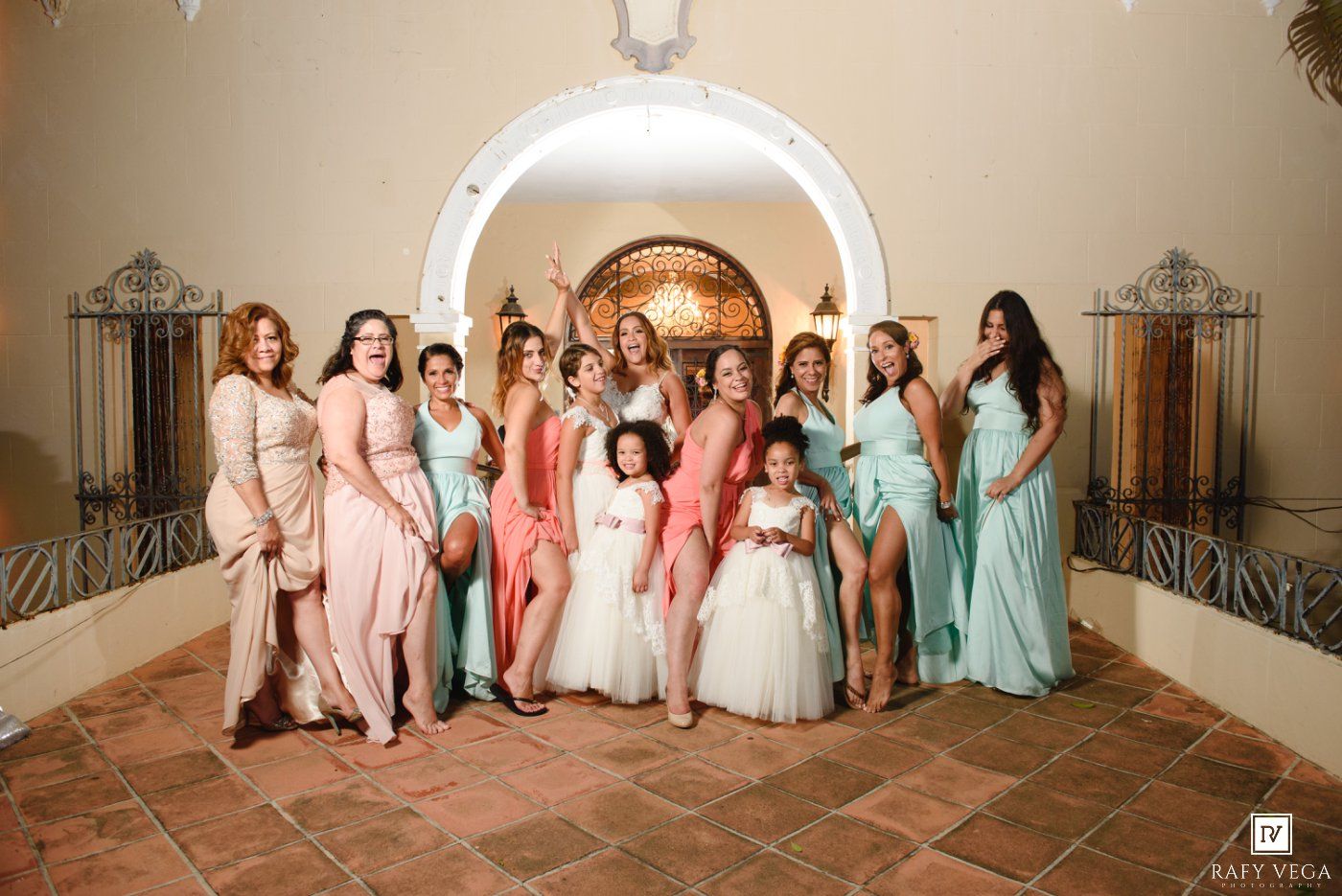 Bride poses with bridesmaids and flower girls in pink and mint dresses under an elegant arched venue.