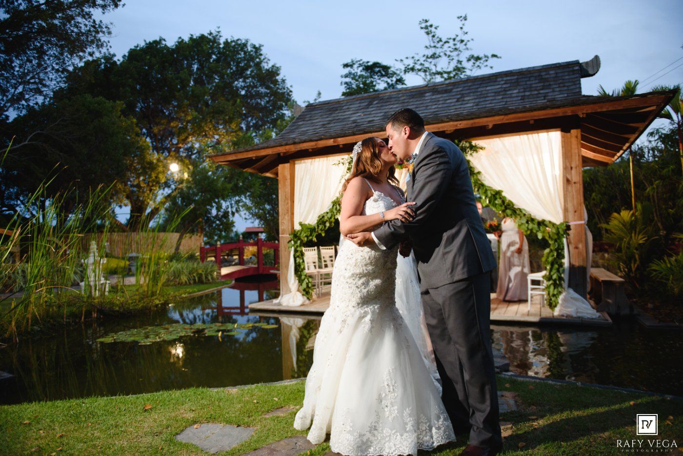 Bride and groom share first dance near a pond with a floral gazebo backdrop at an outdoor garden wedding.