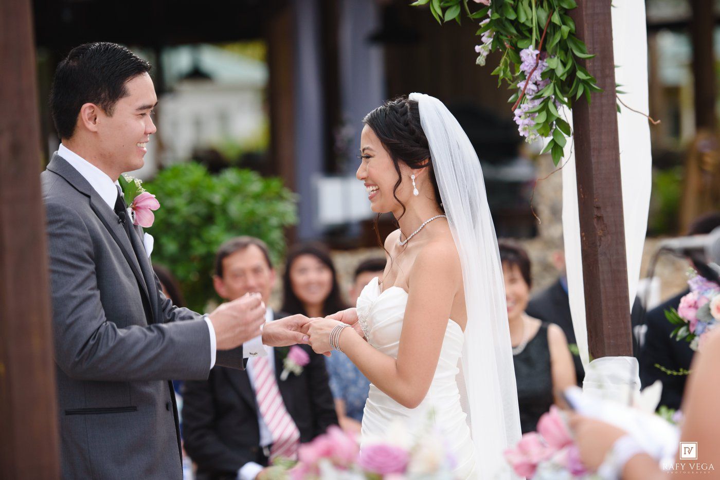Bride and groom exchange rings during an outdoor wedding ceremony with guests seated in the background.