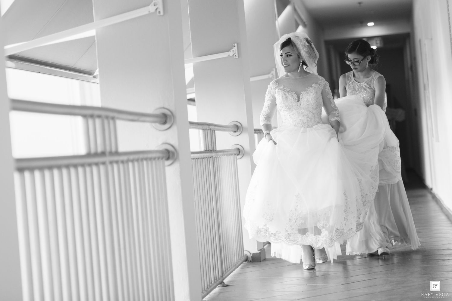 Two women in white wedding gowns walking down a hotel corridor in a black and white wedding photo.