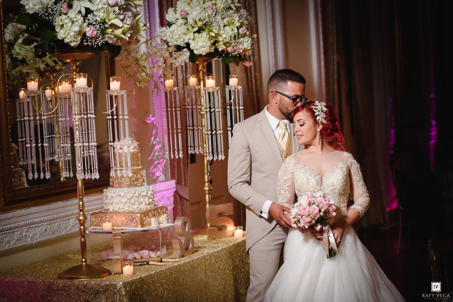 Bride and groom sharing a kiss near a tiered wedding cake at a glamorous candlelit reception venue.