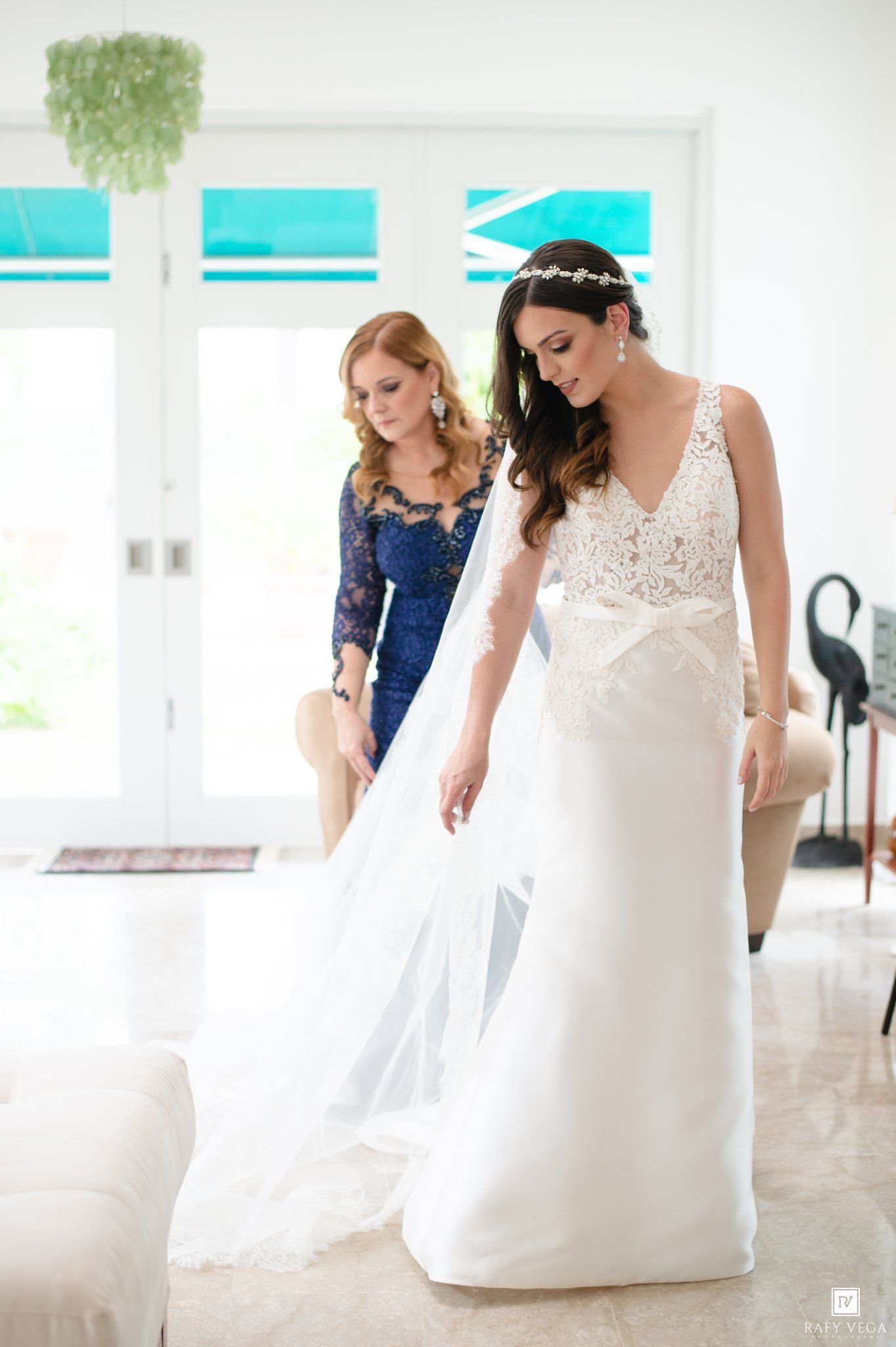 Mother in blue lace dress helps bride adjust white wedding gown train indoors.