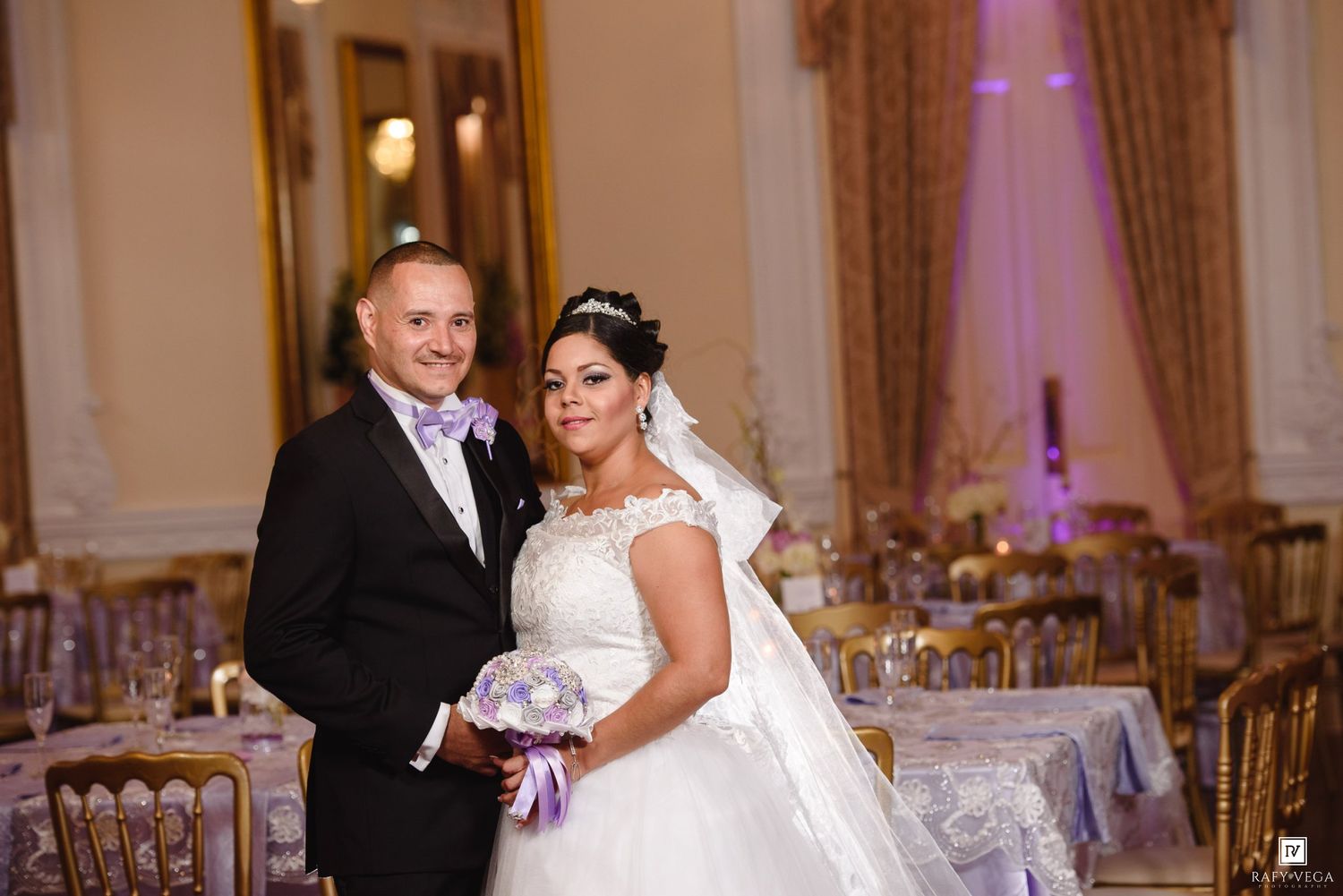 Bride and groom posing together in elegant wedding hall, bride holding purple bouquet in white lace gown.