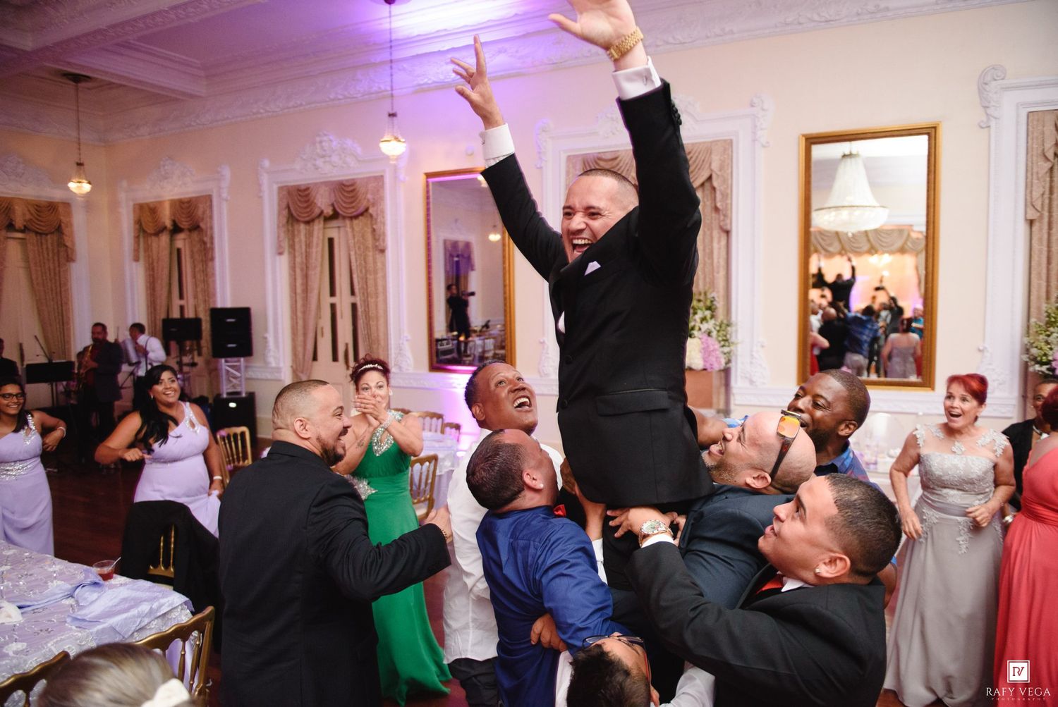 Groom lifted by groomsmen during lively wedding reception celebration in elegant ballroom.