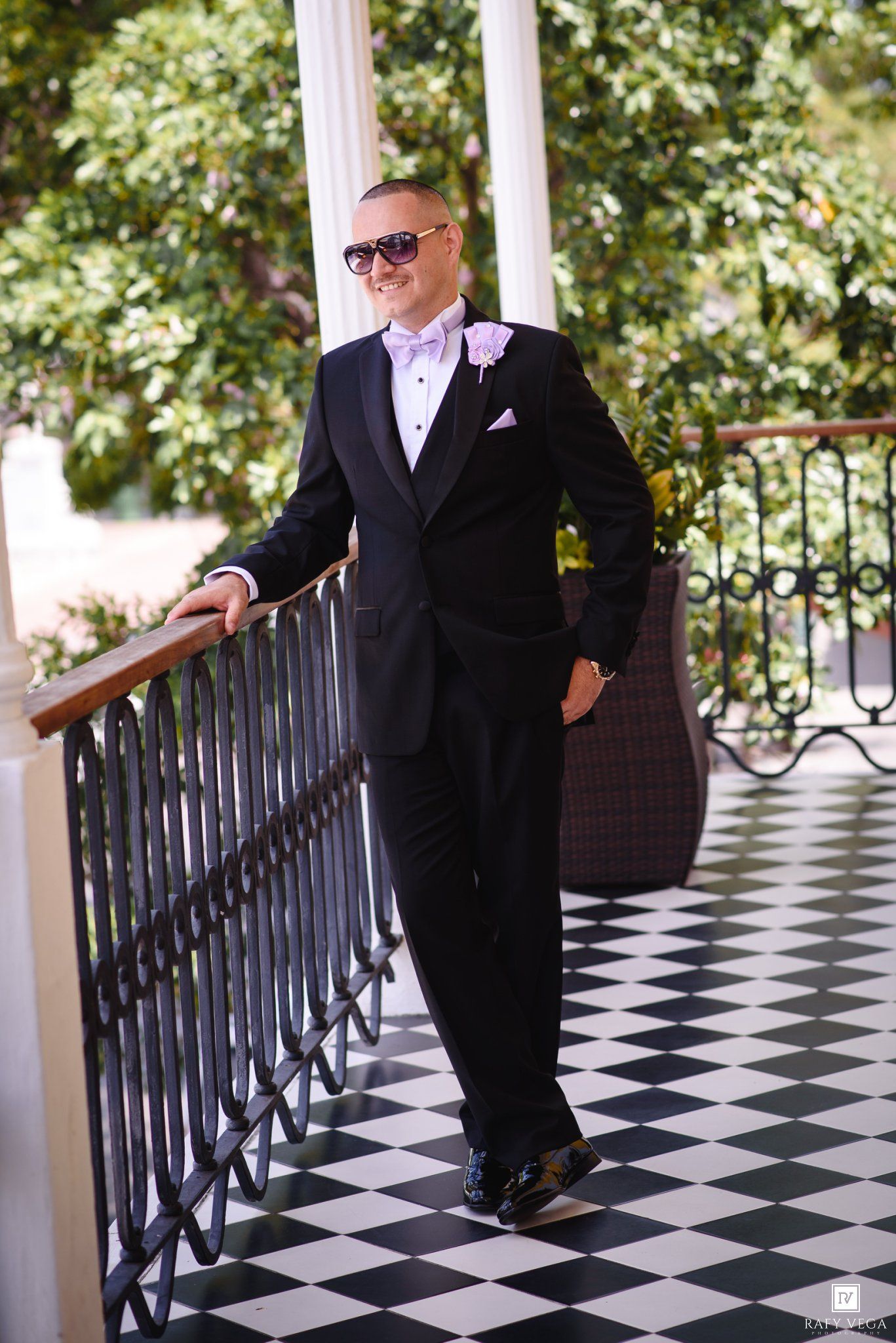Groom in black tuxedo with lavender bow tie posing on checkered balcony wearing sunglasses.