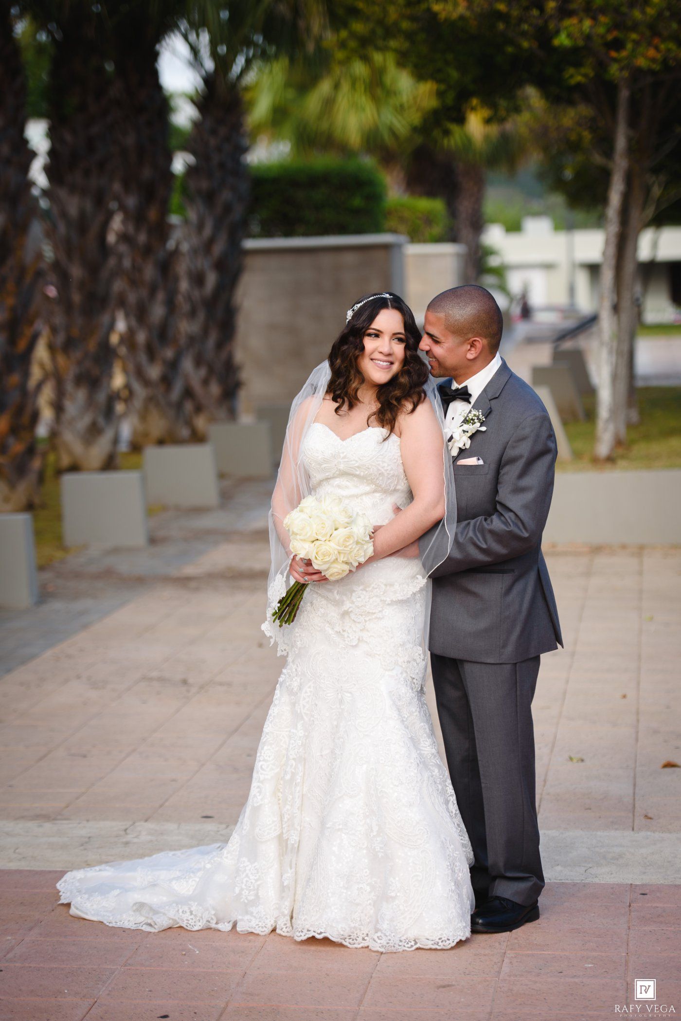 Bride in lace gown holding white bouquet stands with groom in gray tuxedo outdoors near palm trees.