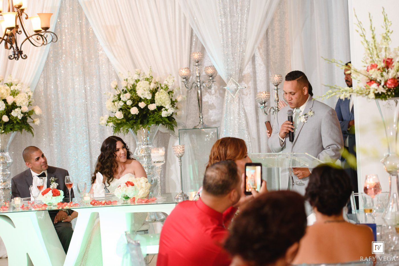 A man in a gray suit gives a speech at an elegant wedding reception with floral arrangements and draped white decor.