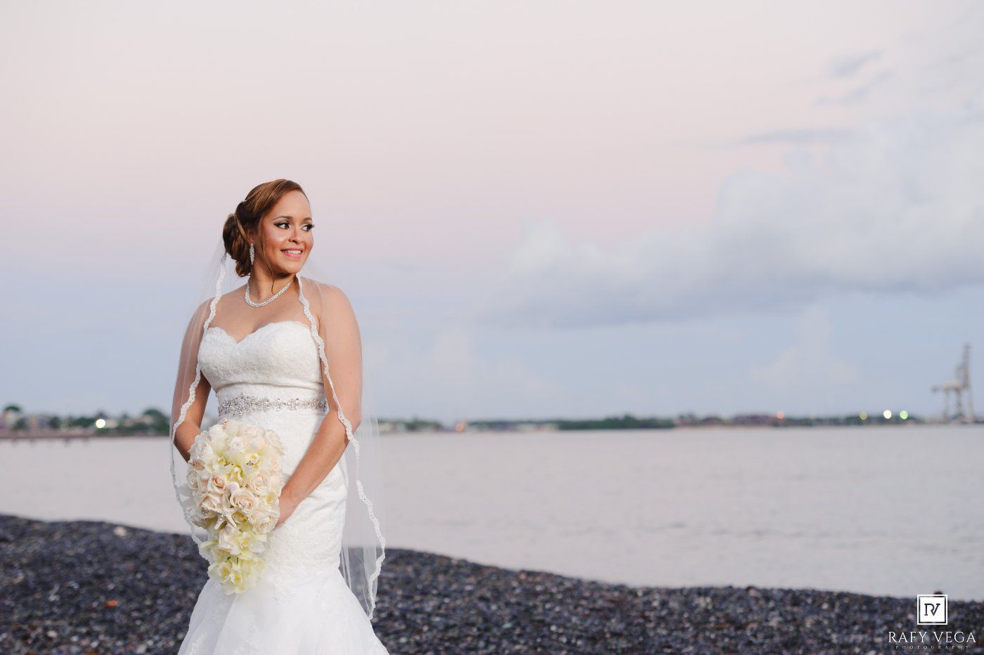 Bride in white halter gown holds bouquet on rocky beach shore at sunset during wedding portrait.