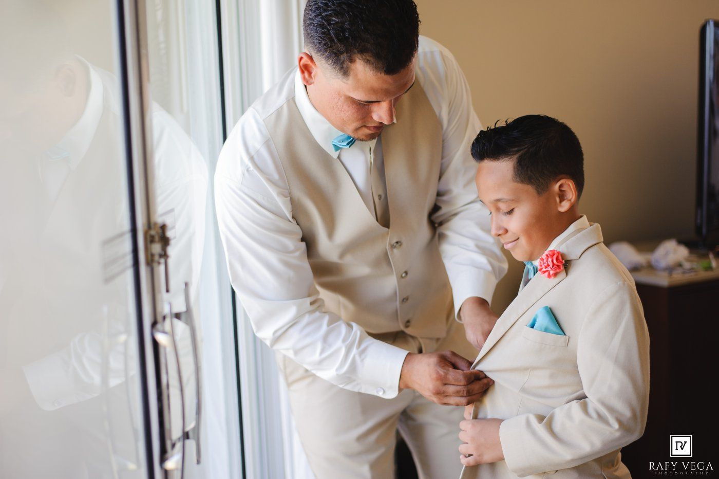 A man in a beige suit adjusts the jacket of a young boy in a matching suit with a coral boutonniere.