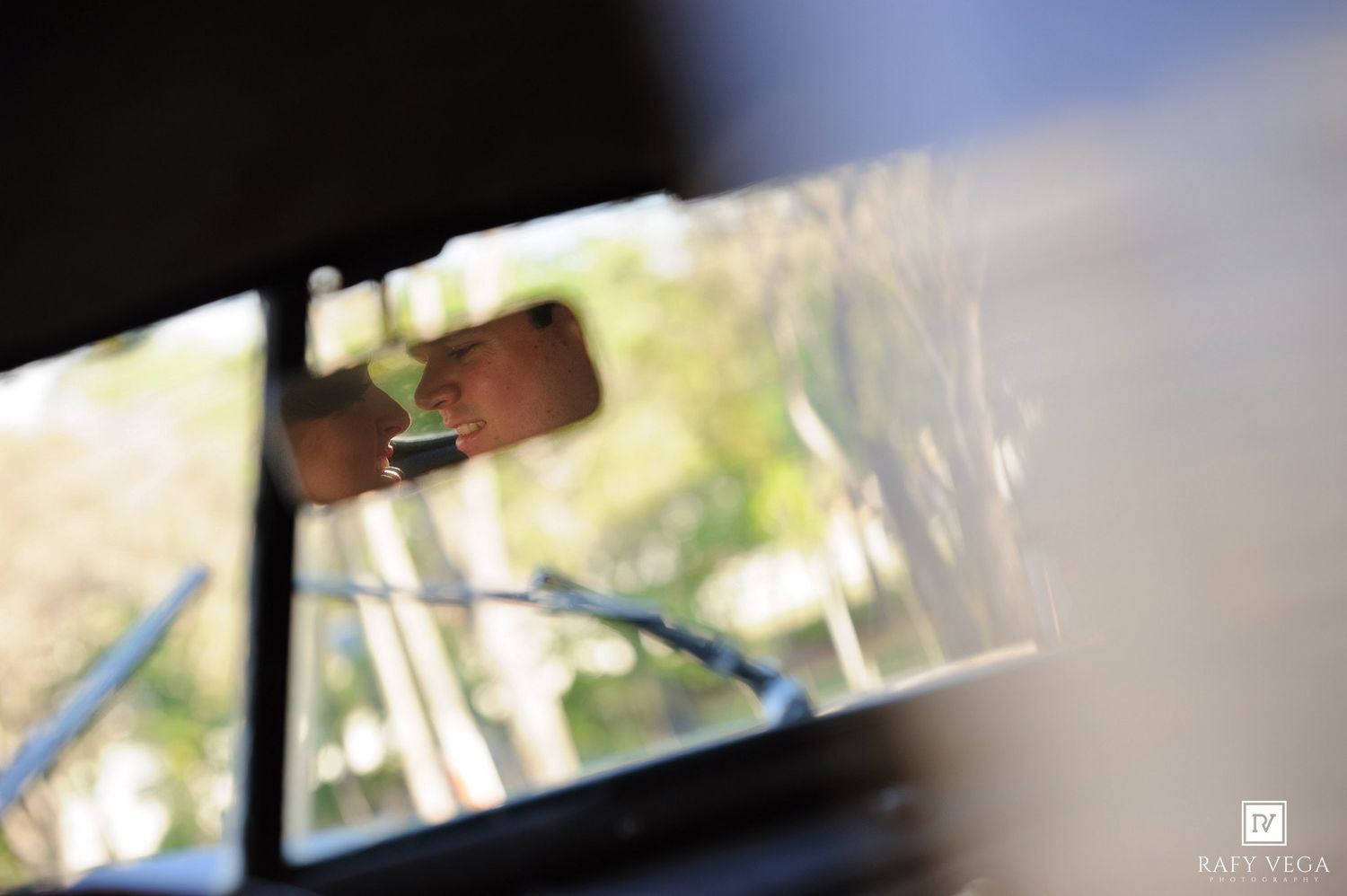 Reflection of a person in a car rearview mirror surrounded by lush green trees on a sunny day.