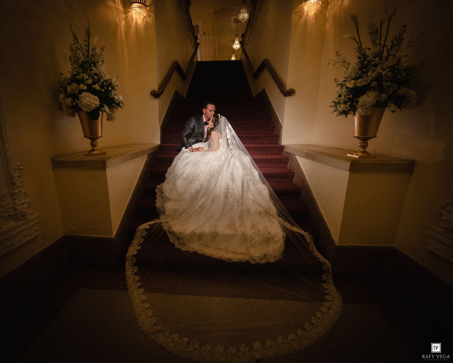 Bride in a stunning ballgown and groom pose on a grand staircase with floral arrangements at their wedding venue.