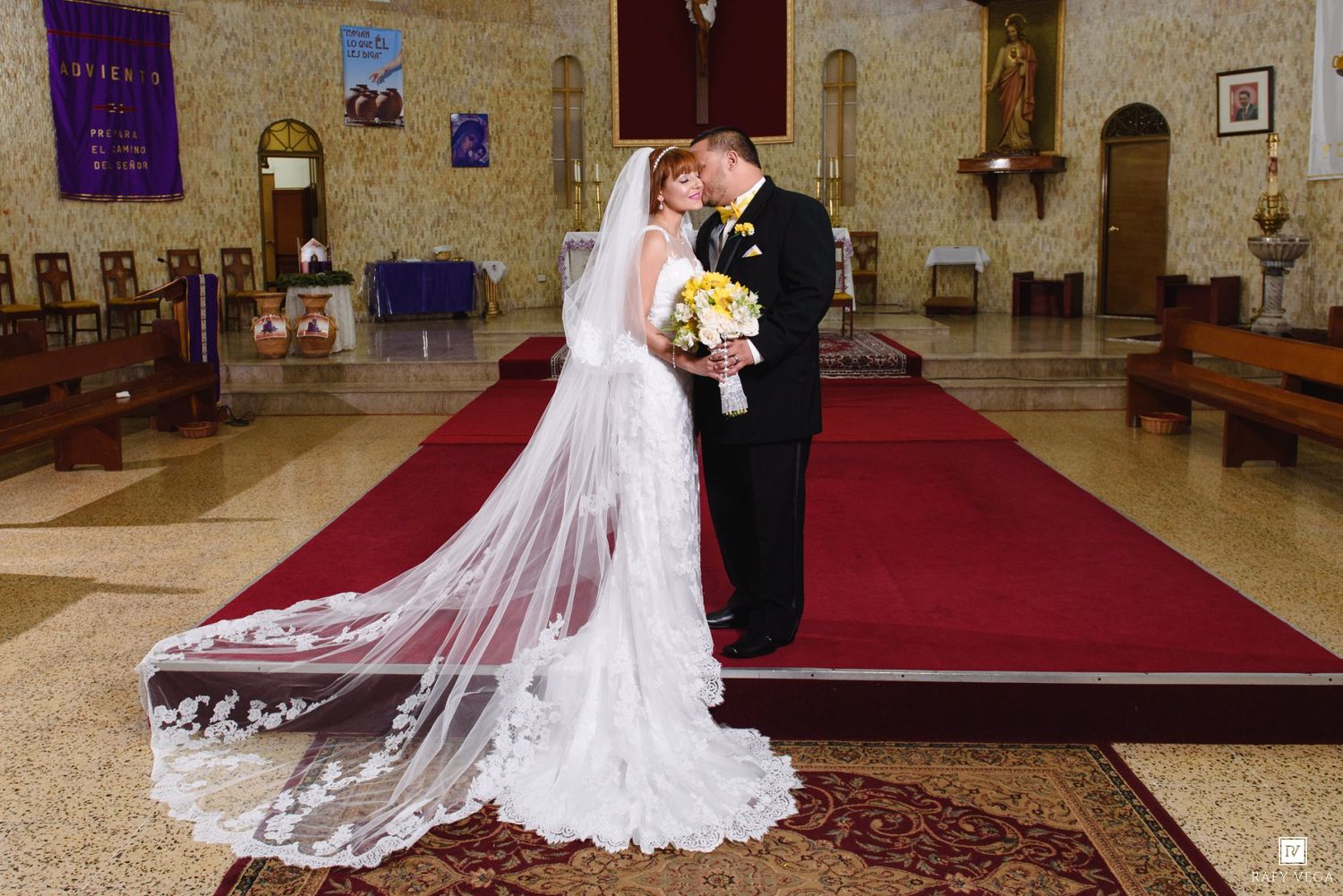 Bride and groom share a romantic kiss at the altar of an elegant church with red carpet aisle on their wedding day.