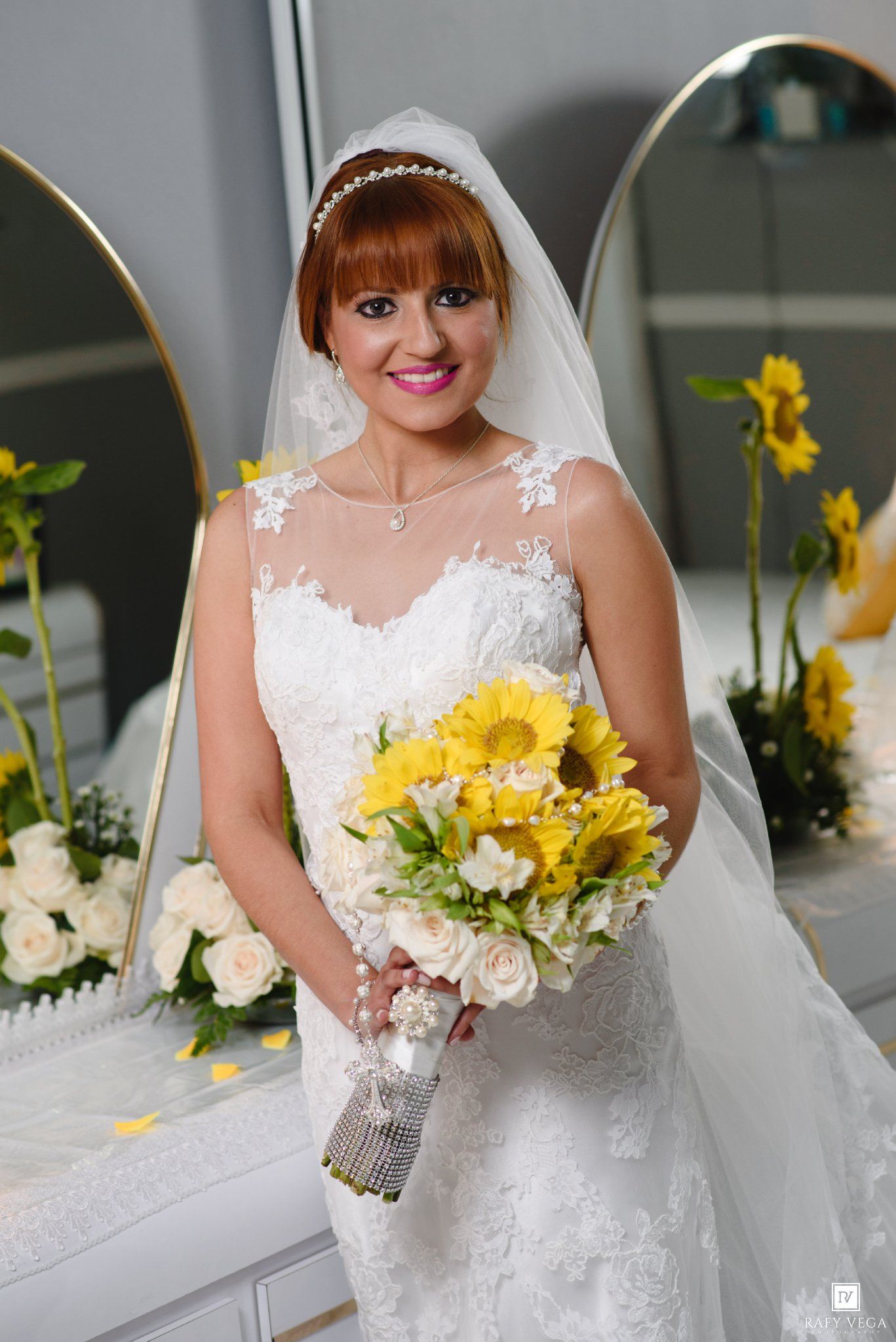 Bride in lace wedding gown holding yellow sunflower bouquet poses near mirrors before ceremony.