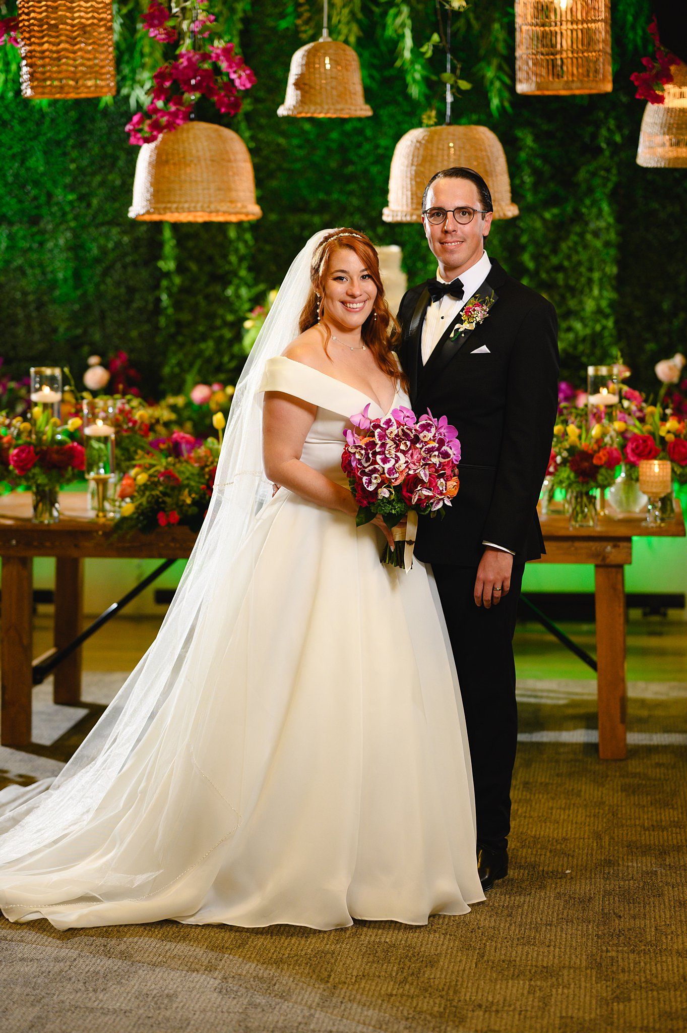 Bride in off-shoulder white ball gown holding pink bouquet stands with groom in black tuxedo at floral wedding reception.