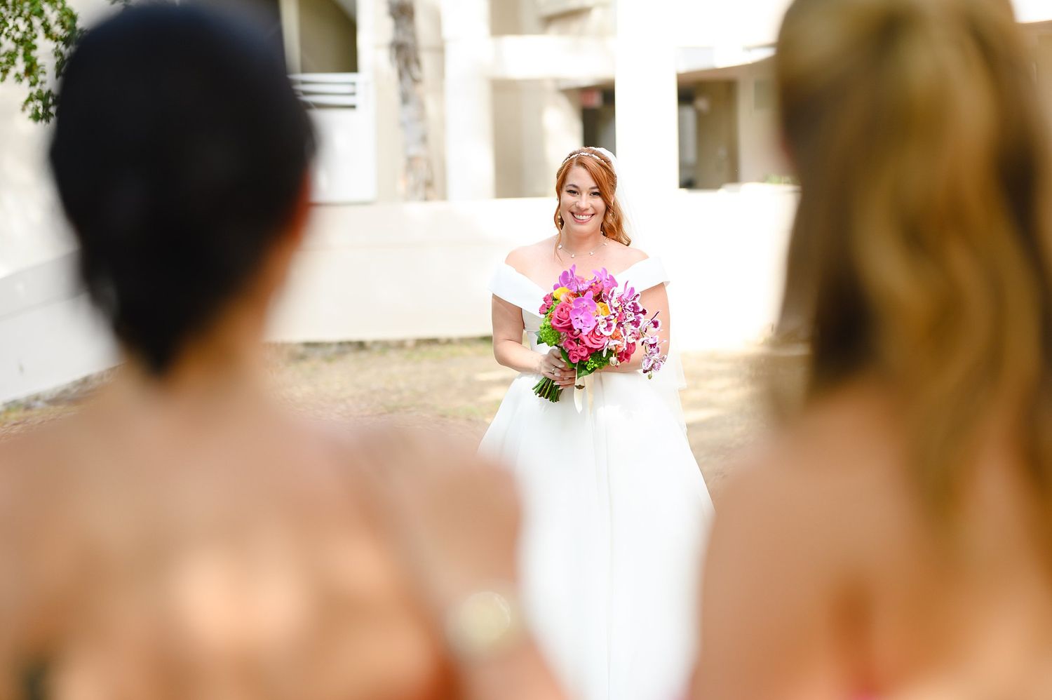 Smiling bride in white gown holds vibrant pink bouquet while bridesmaids in coral dresses look on at outdoor wedding.
