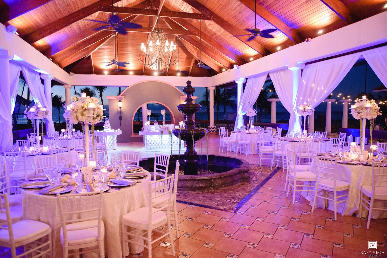Elegant wedding reception venue with white chairs, floral centerpieces, fountain, and purple ambient lighting under vaulted ceiling.