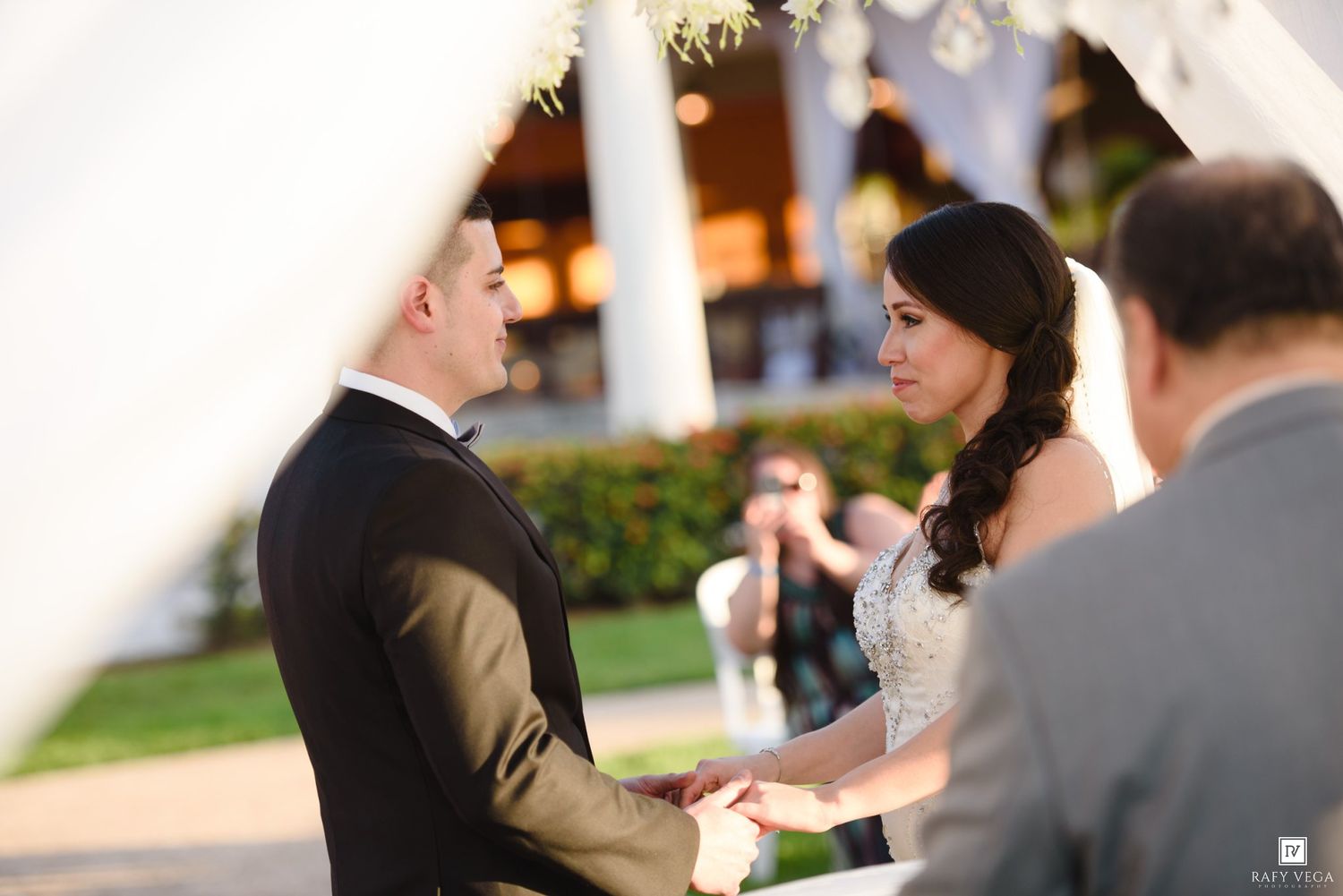 Bride and groom exchange vows during an outdoor wedding ceremony, holding hands before an officiant.