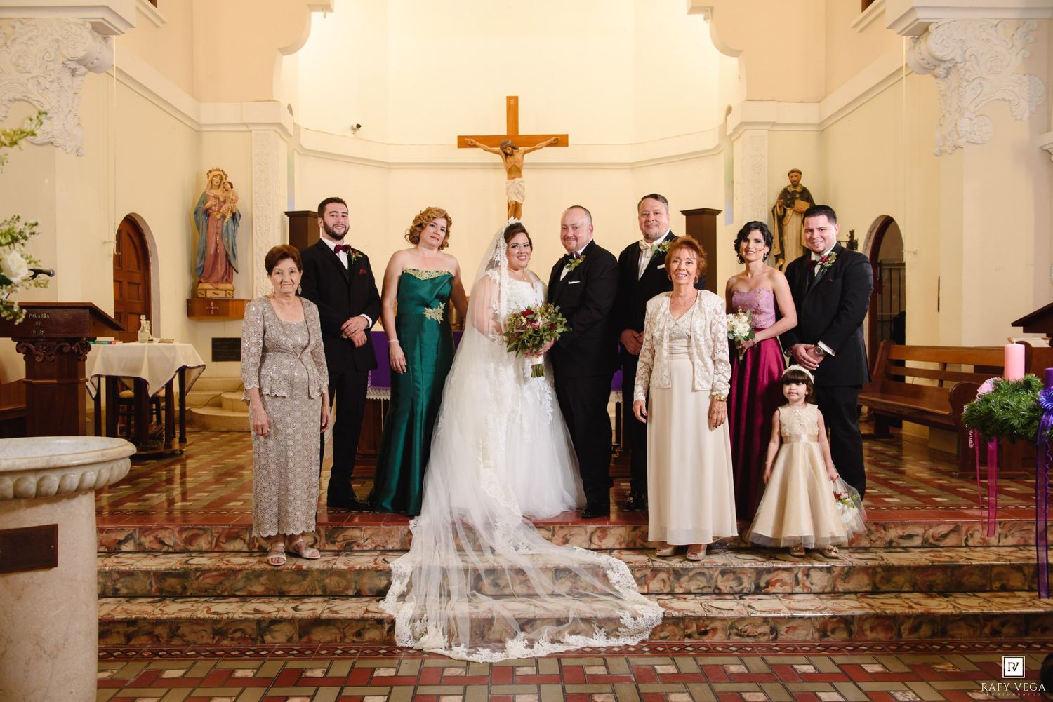 Wedding party poses in a church with a bride in white gown, groom, and family members in elegant formal attire.