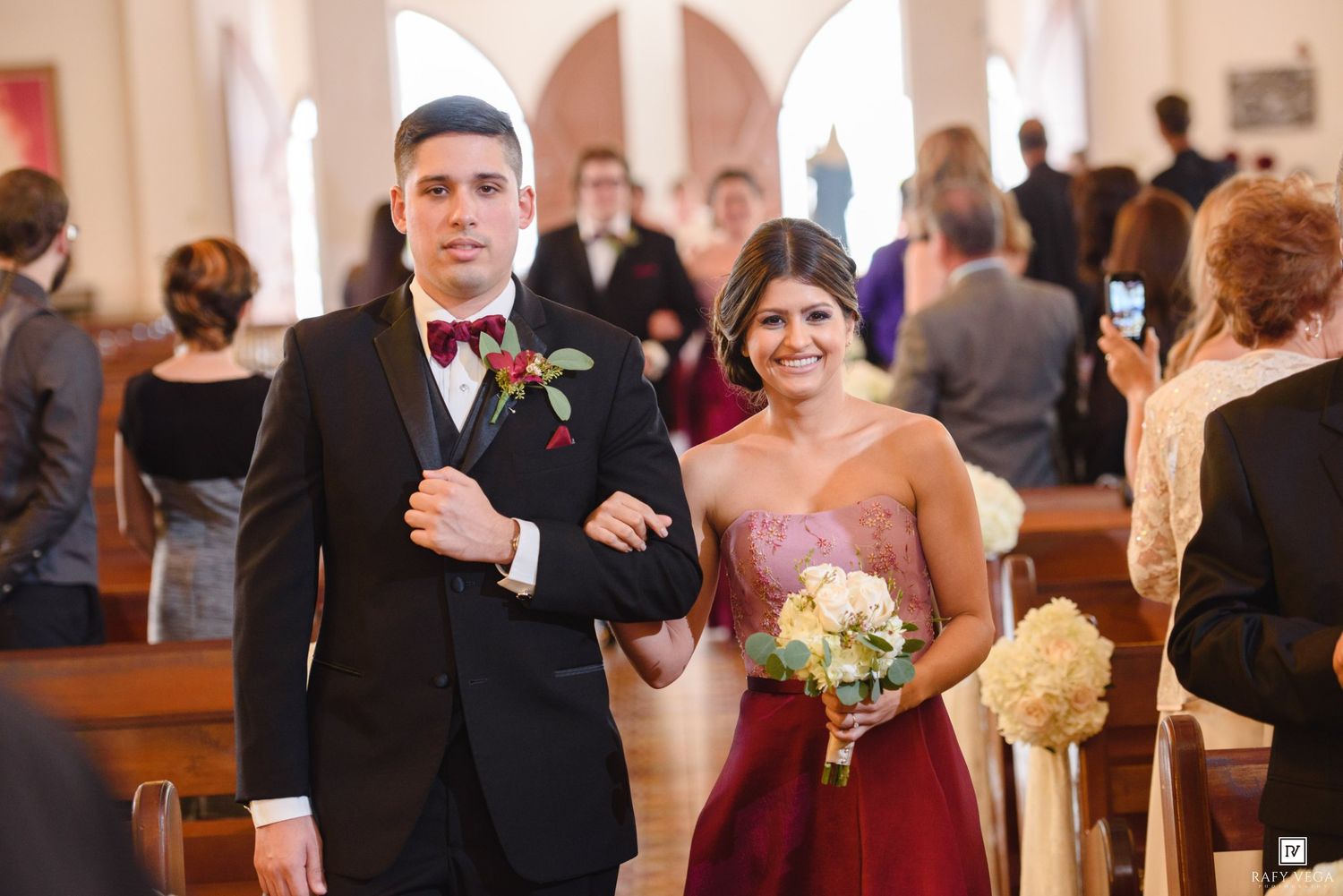 Groomsman in black tuxedo escorts bridesmaid in burgundy gown down church aisle during wedding ceremony.