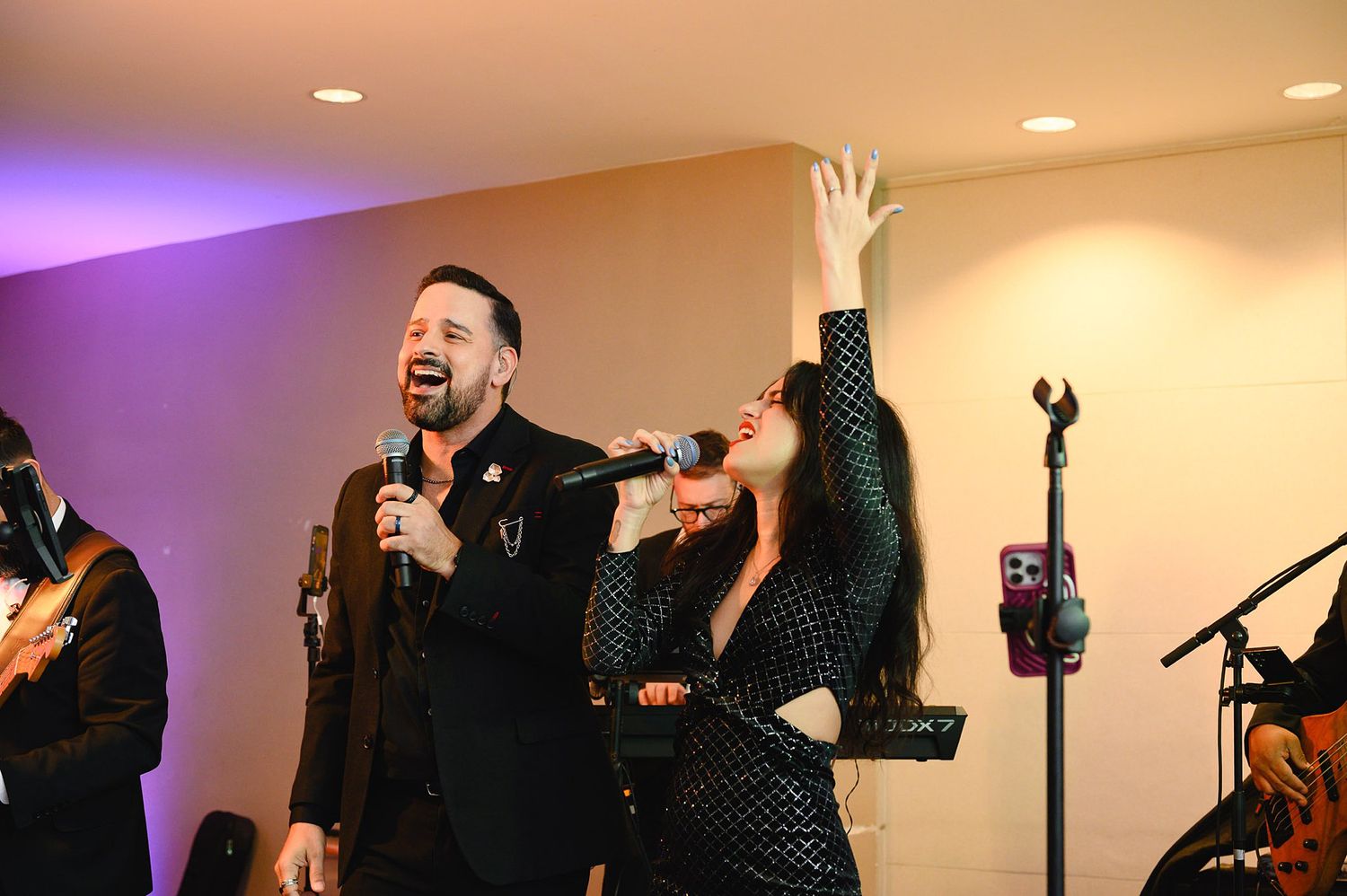 Two singers performing on stage, man in black suit holding mic, woman in sparkly dress reaching up dramatically.