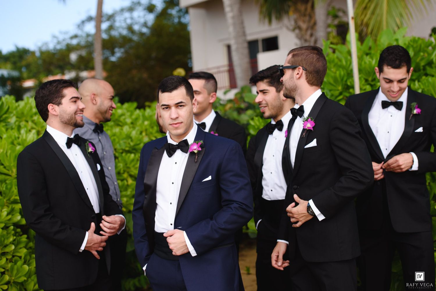 Groom and groomsmen in black tuxedos with pink boutonnieres posing outdoors at a wedding celebration.