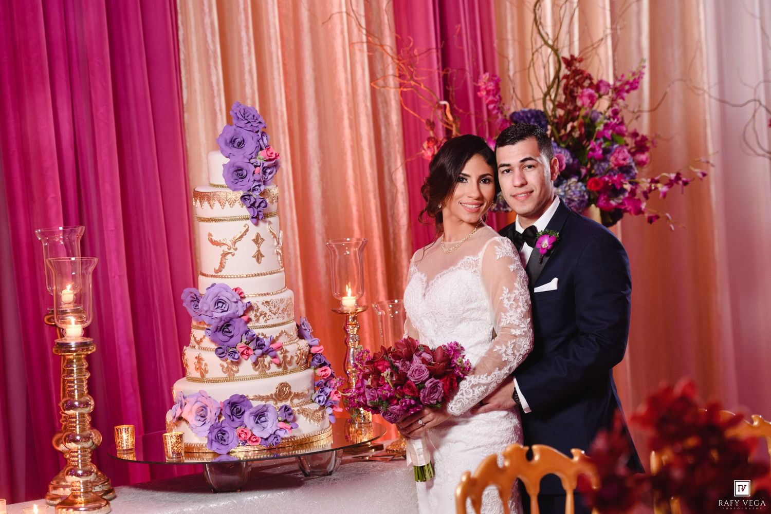 Bride and groom pose beside elegant multi-tier wedding cake with purple flowers at romantic pink-draped reception.