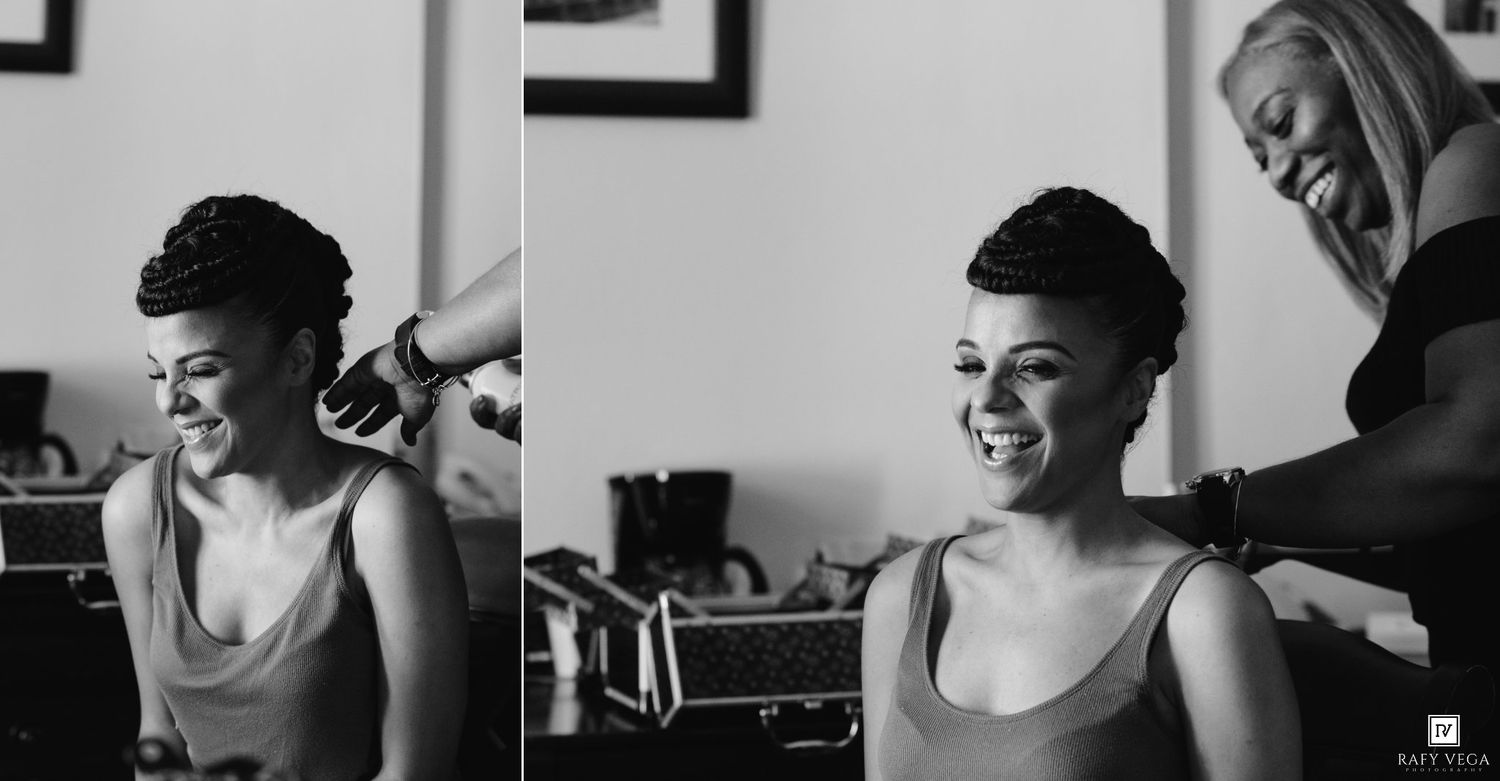 Black and white photo of a bride smiling joyfully as her hairstylist works on her elegant updo hairstyle before her wedding.