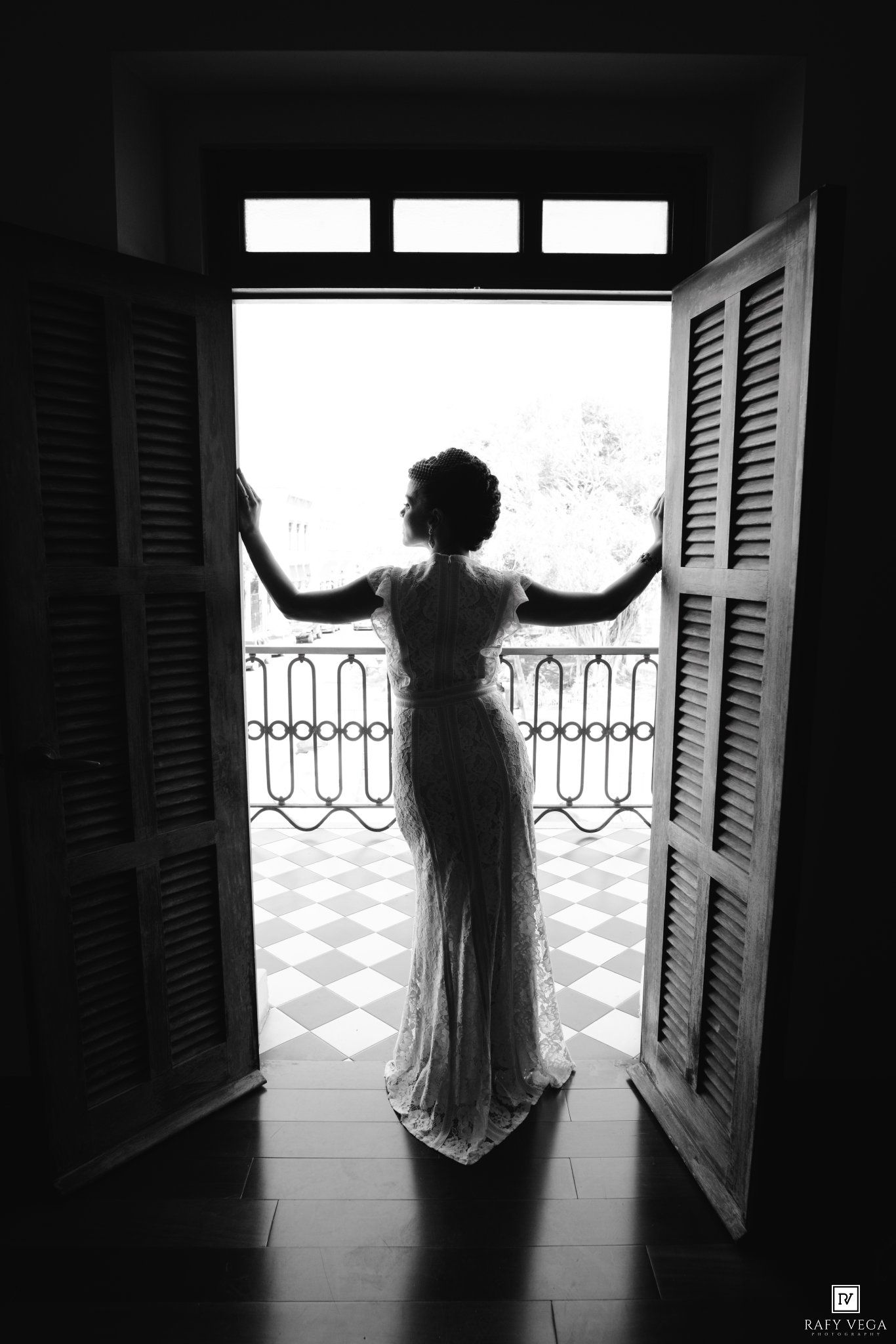 Black and white photo of a bride in a white gown standing in open French doors leading to a balcony.