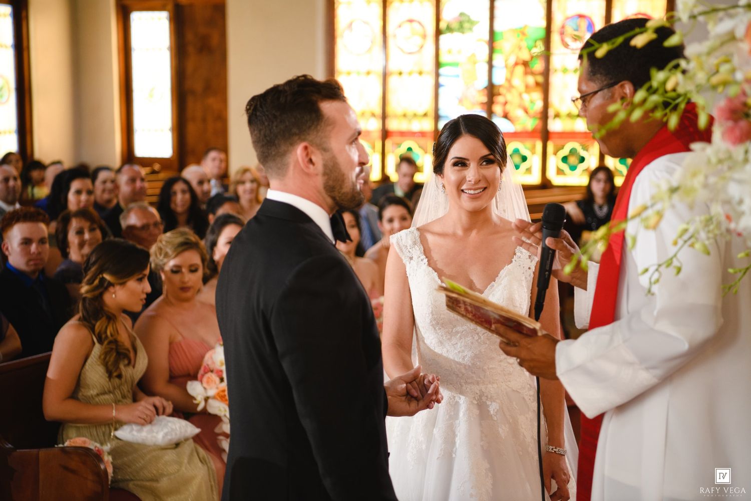Bride and groom exchange vows at church altar with officiant beside floral arrangements during wedding ceremony.