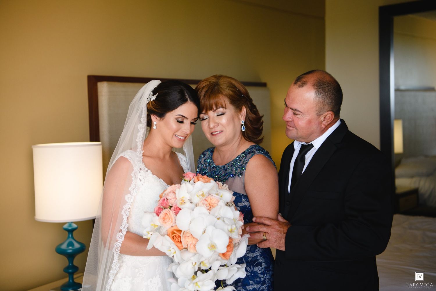 Bride in white lace gown and veil holds bouquet while sharing emotional moment with parents in hotel room.