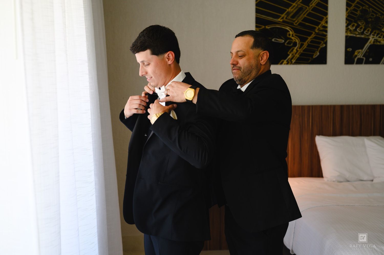 Two men in black suits preparing for a wedding, one helping the other adjust his tie in a hotel room.