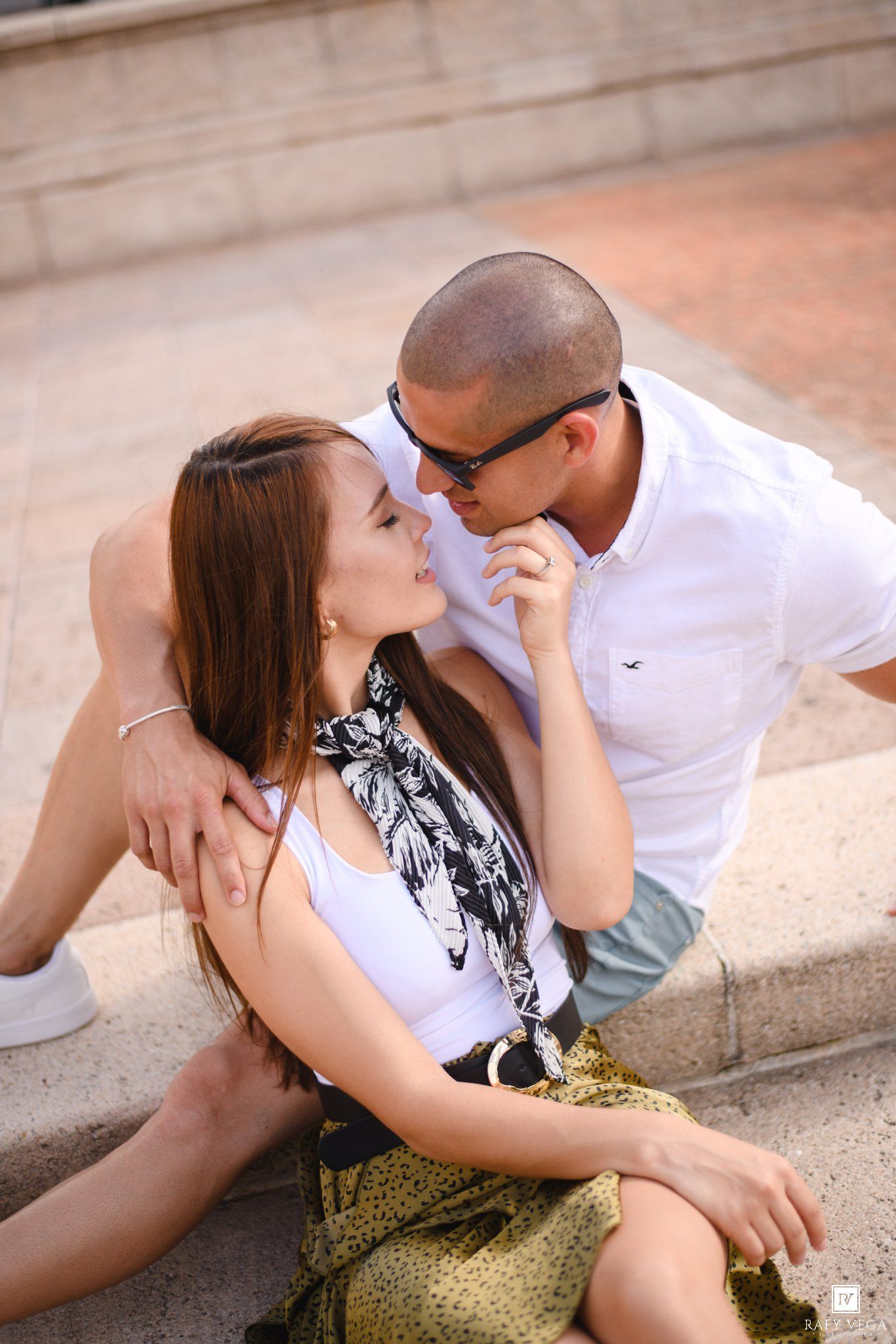 Couple sharing a romantic moment on stone steps, woman in white tank top and floral scarf, man in white polo with sunglasses.