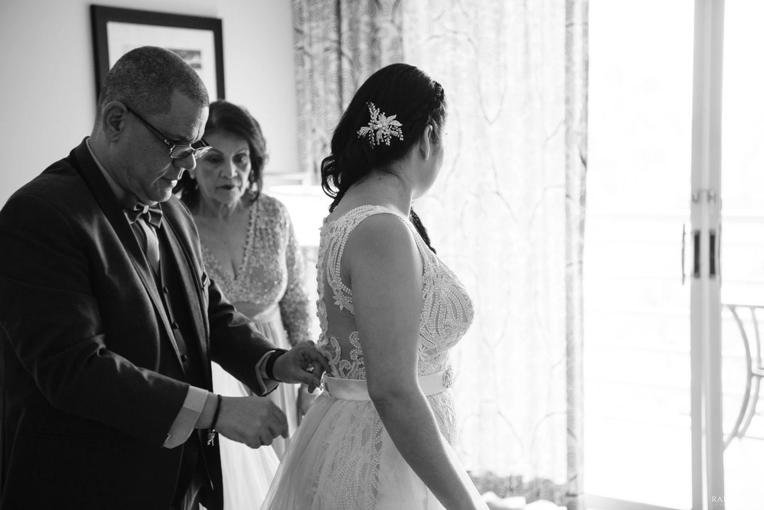 Black and white wedding photo of bride with family helping her prepare near a bright window.