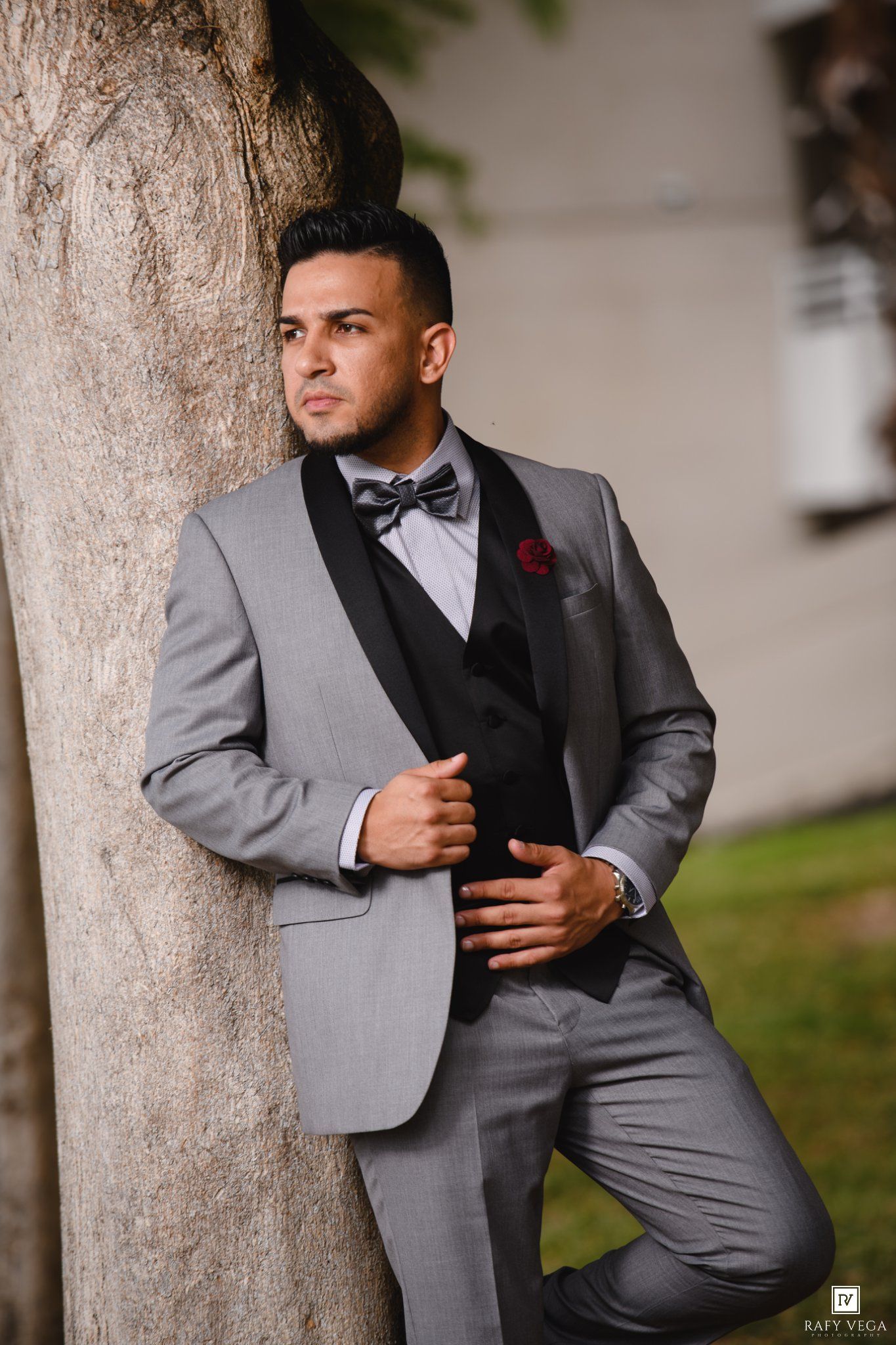 Elegant groom in gray tuxedo with black lapels and bow tie leaning against tree outdoors.