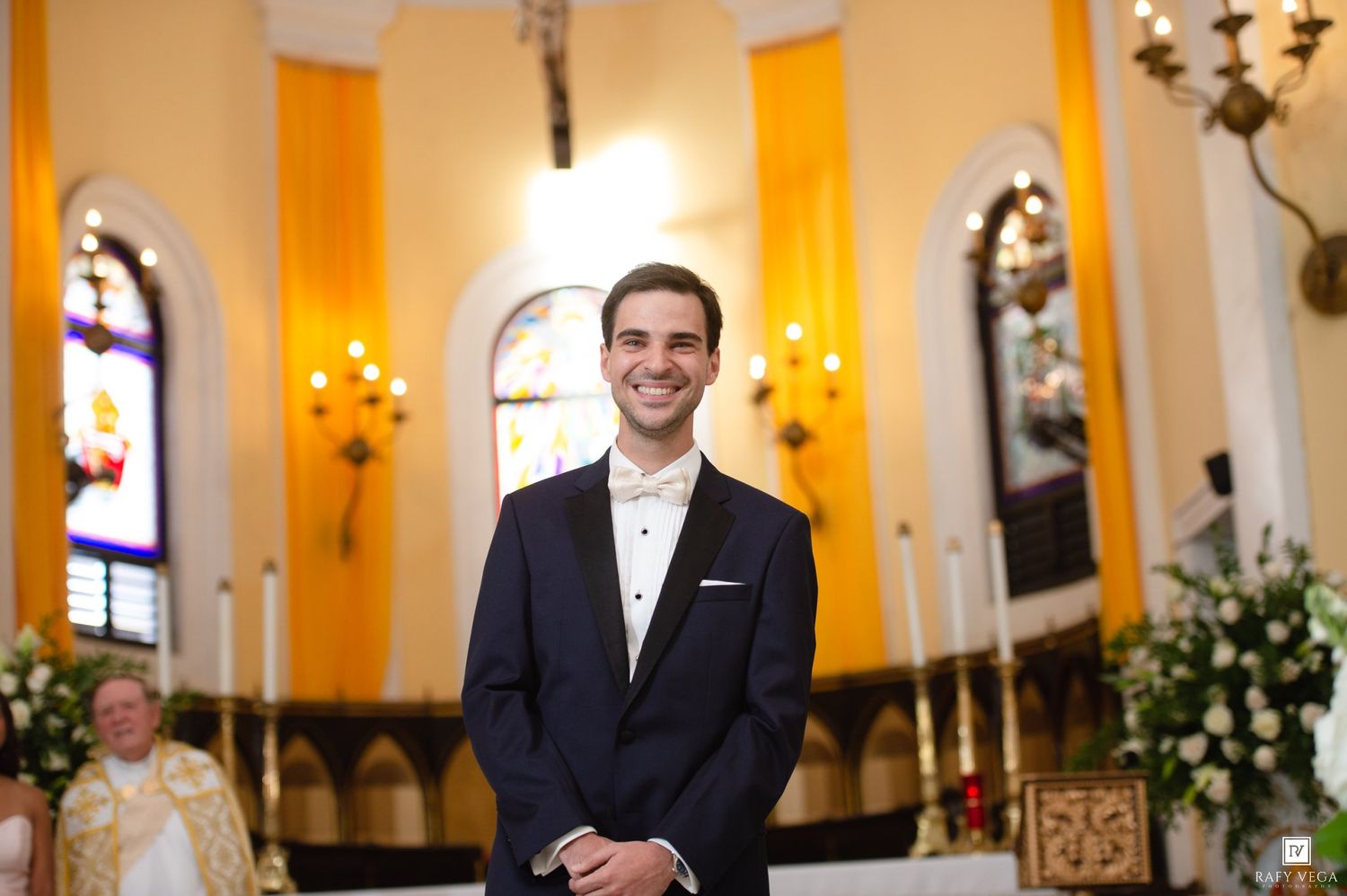 Smiling groom in navy tuxedo stands at altar of ornate golden church.