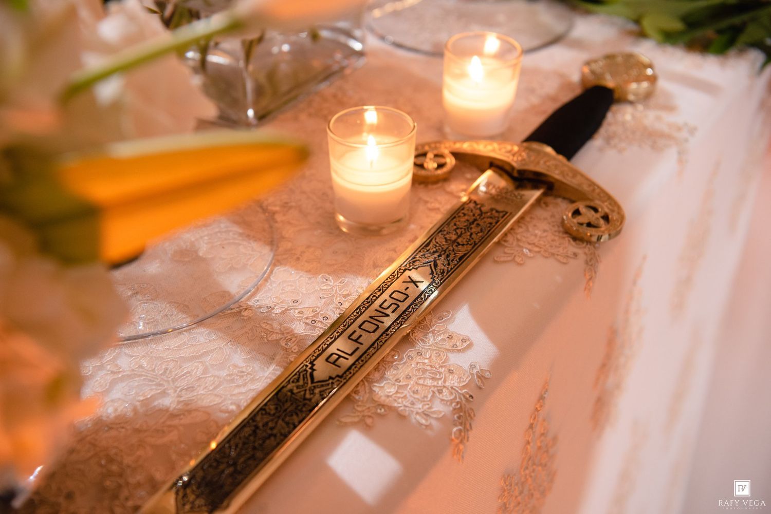 Ornate gold sword with engraving displayed on white lace tablecloth beside glowing candles at wedding reception.